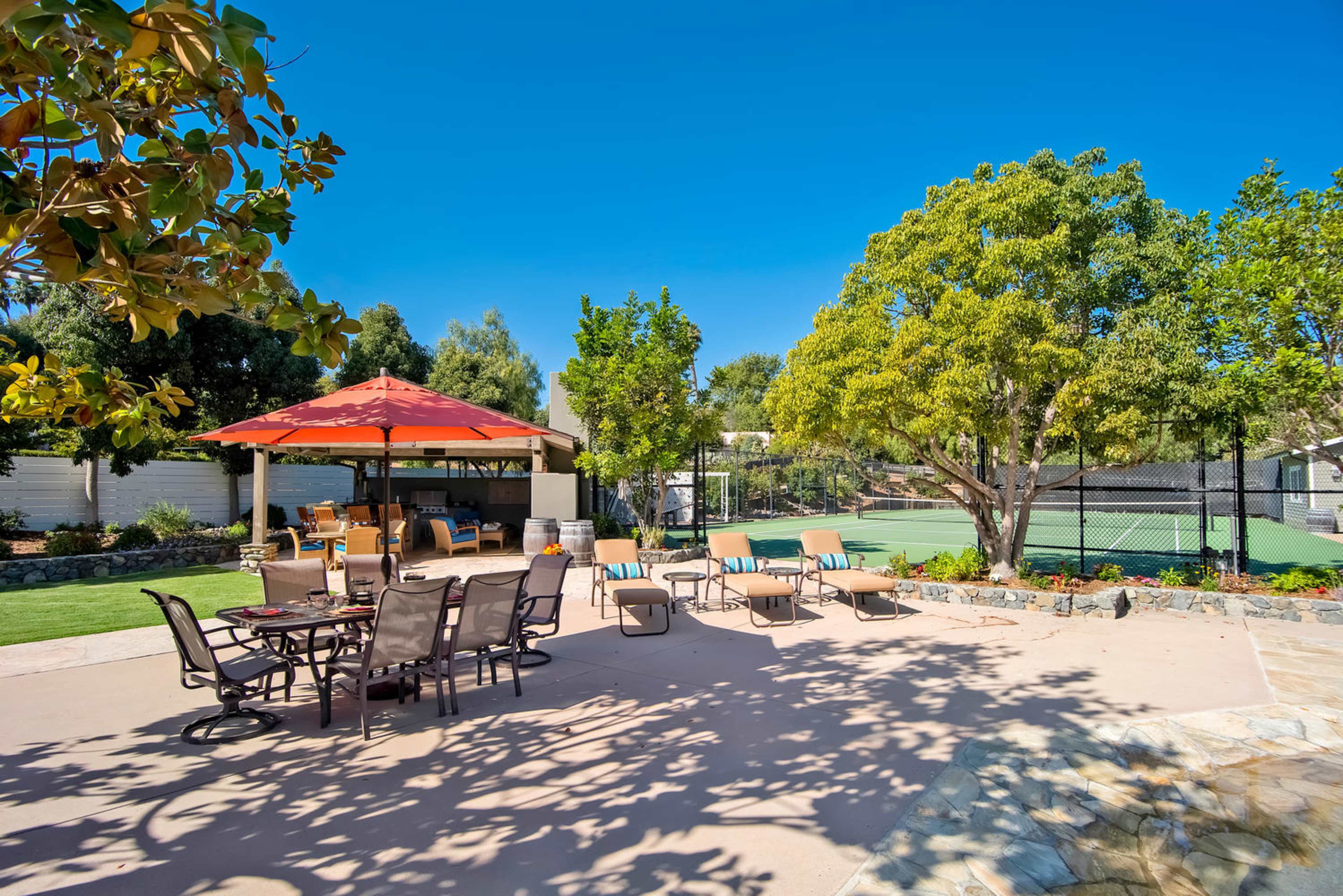 The image shows a sunny backyard area with seating around a shaded table and a tennis court in the background.