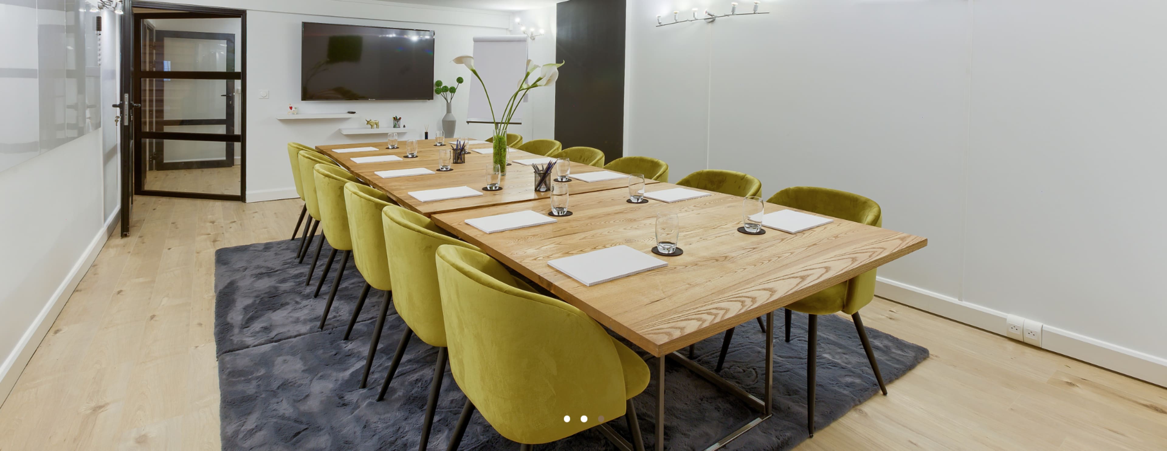 A long wooden conference table with yellow chairs is set up in a modern meeting room featuring a large screen and a wall display.