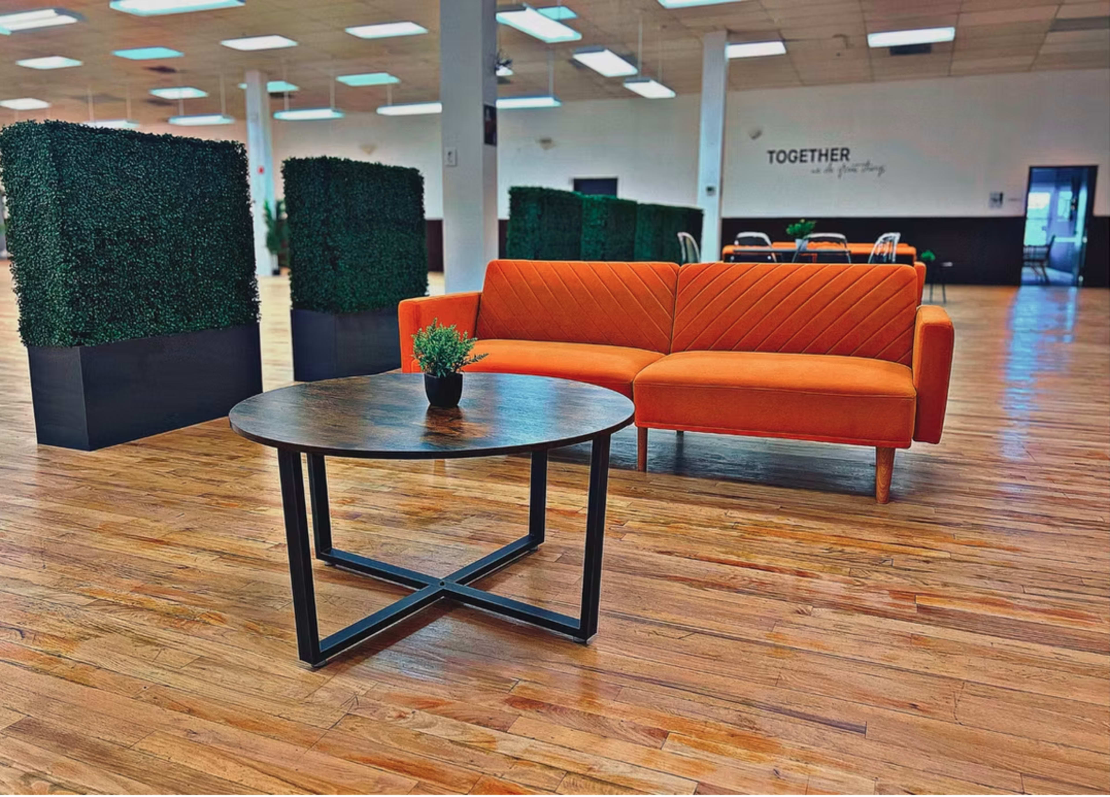 The image shows a modern interior space featuring an orange couch and a round wooden coffee table surrounded by greenery in planters.