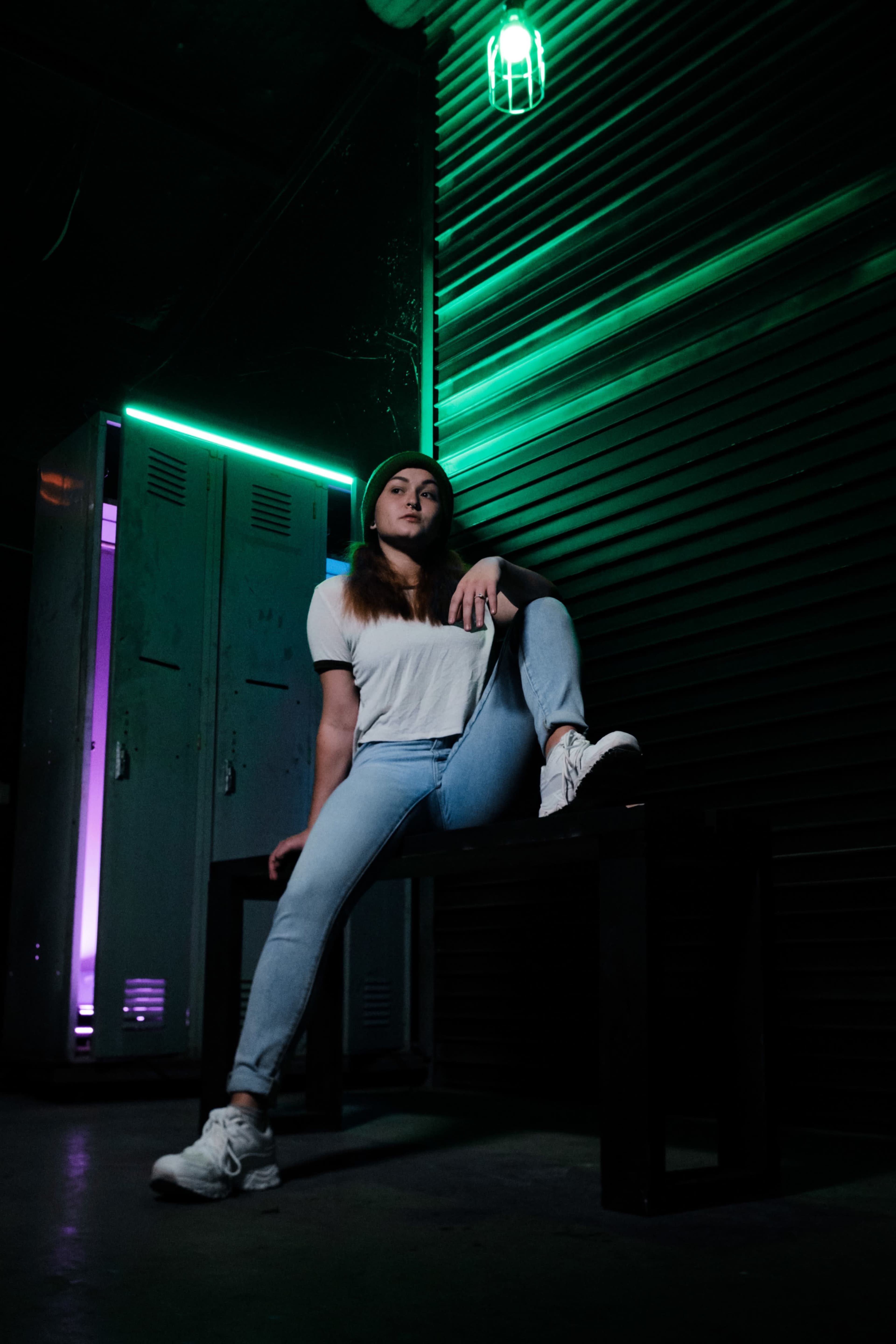 A person sits on a bench in a dimly lit room, surrounded by green and purple lighting, with lockers in the background.