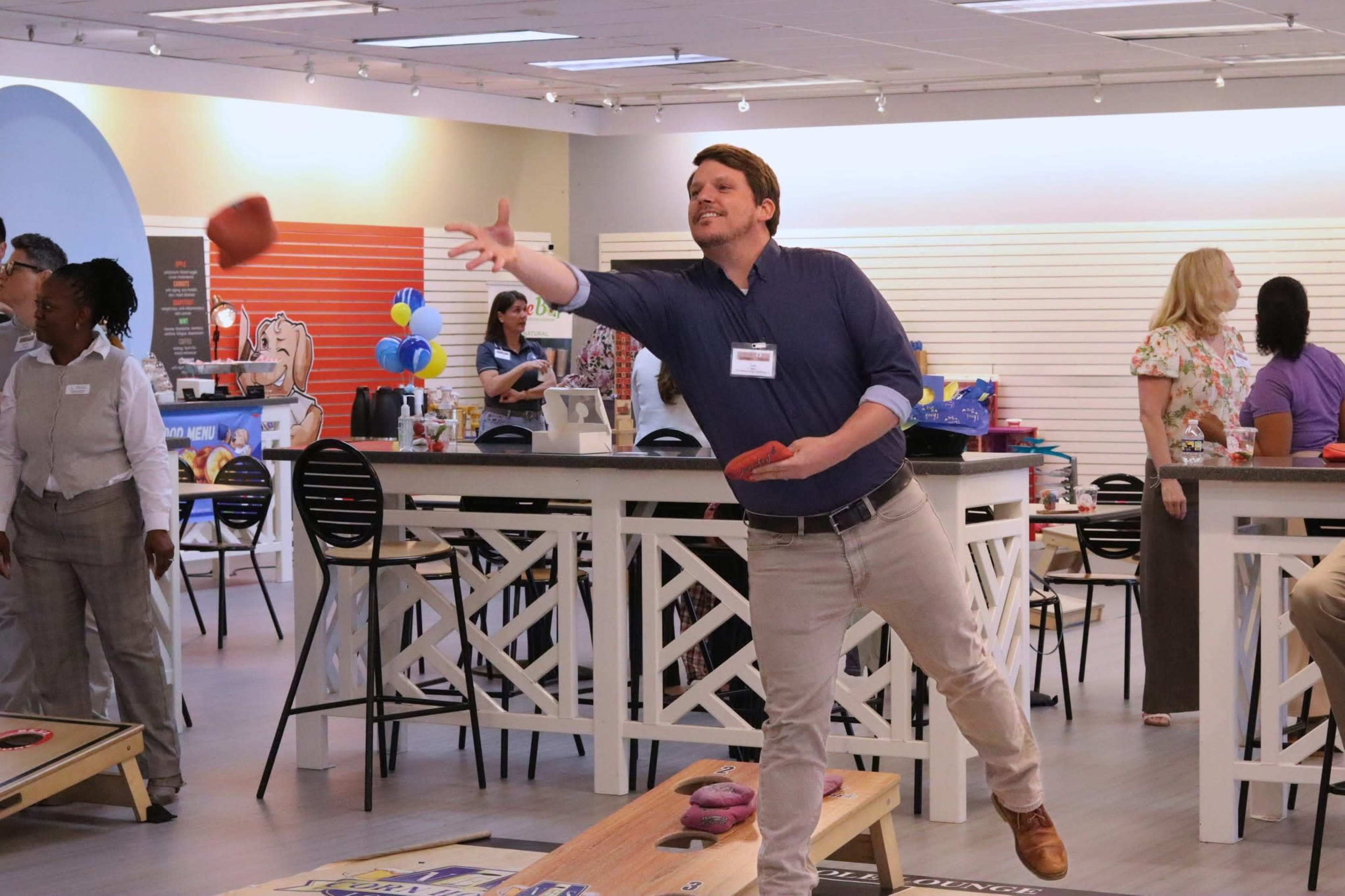 A man wearing a blue shirt is throwing a bean bag towards a cornhole board in a brightly lit event space filled with people.