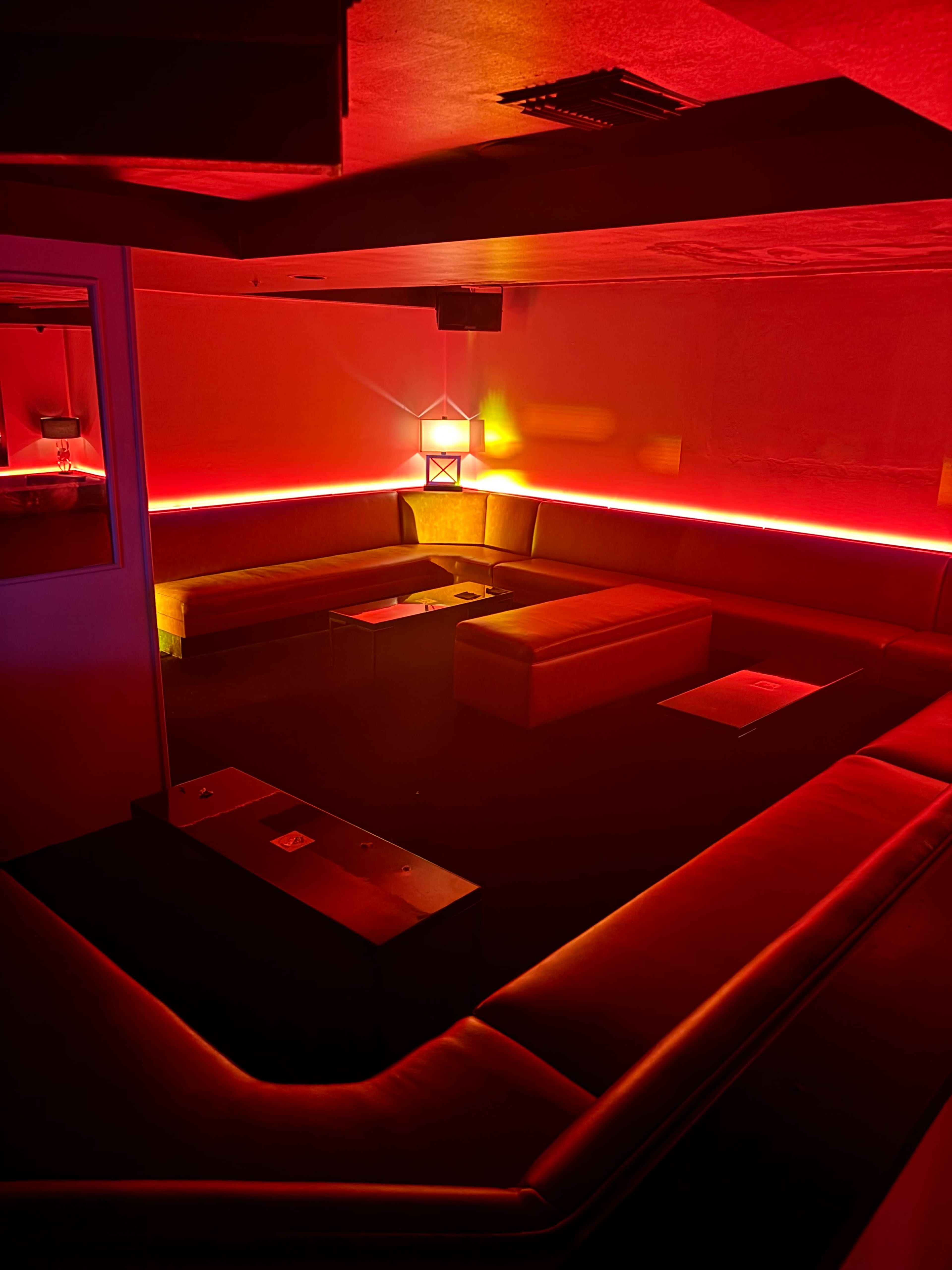 The image shows a dimly lit lounge with red lighting, featuring sofas arranged in a circular layout and low tables.