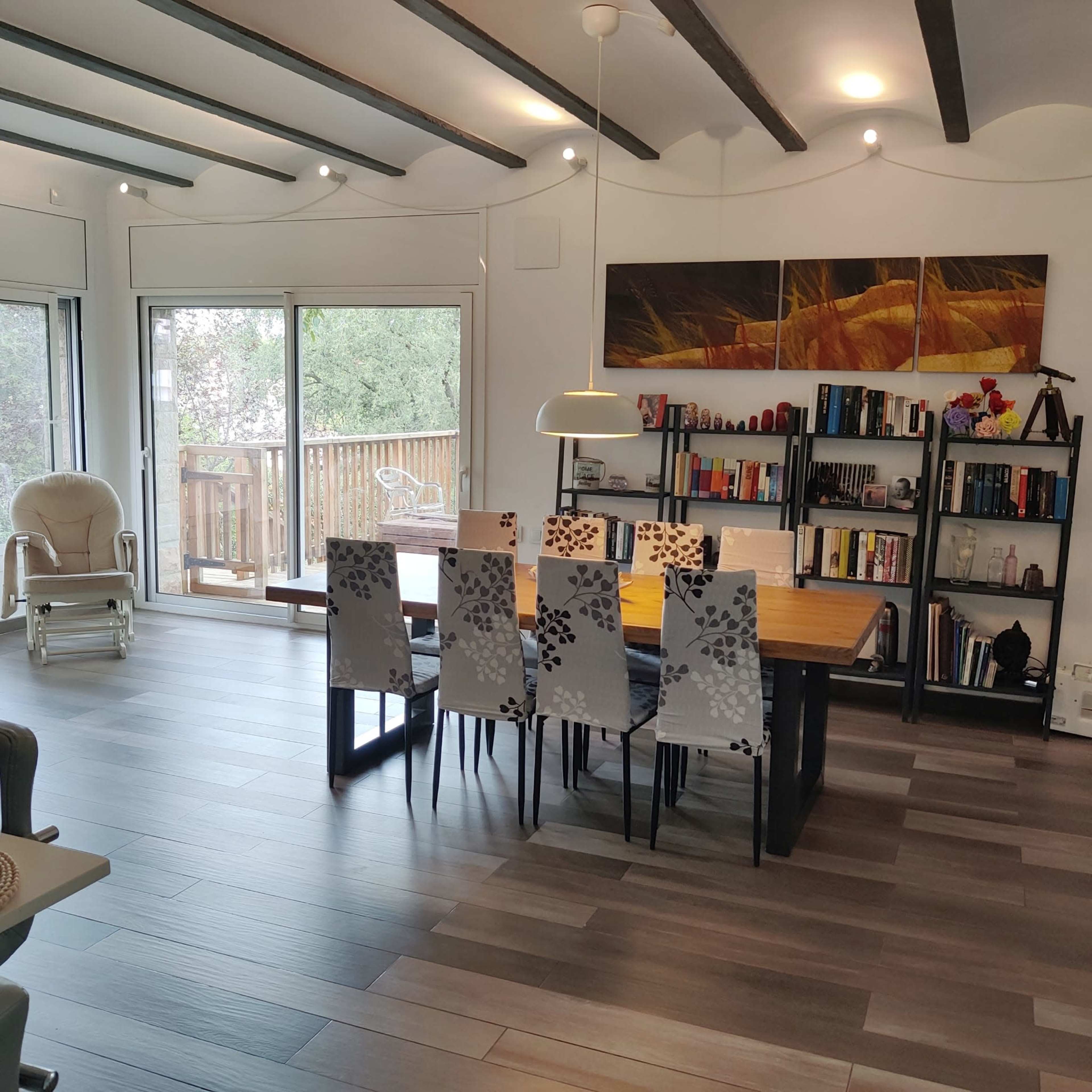 A dining area features a wooden table surrounded by six chairs with decorative backs, set against a backdrop of a large window and bookshelves.