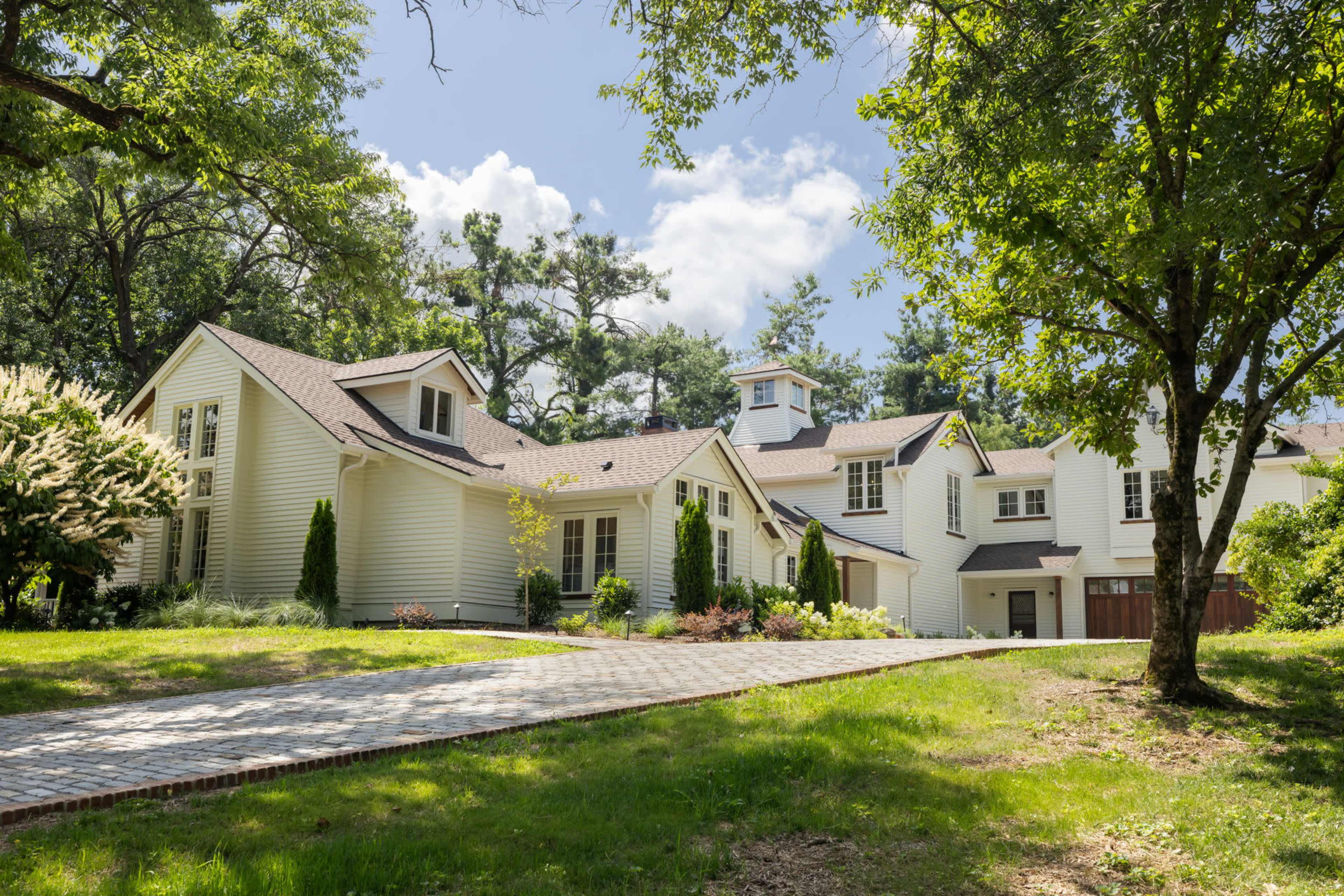 A spacious, modern home with a light-colored exterior is nestled among green trees and a well-kept lawn, with a stone driveway leading up to it.