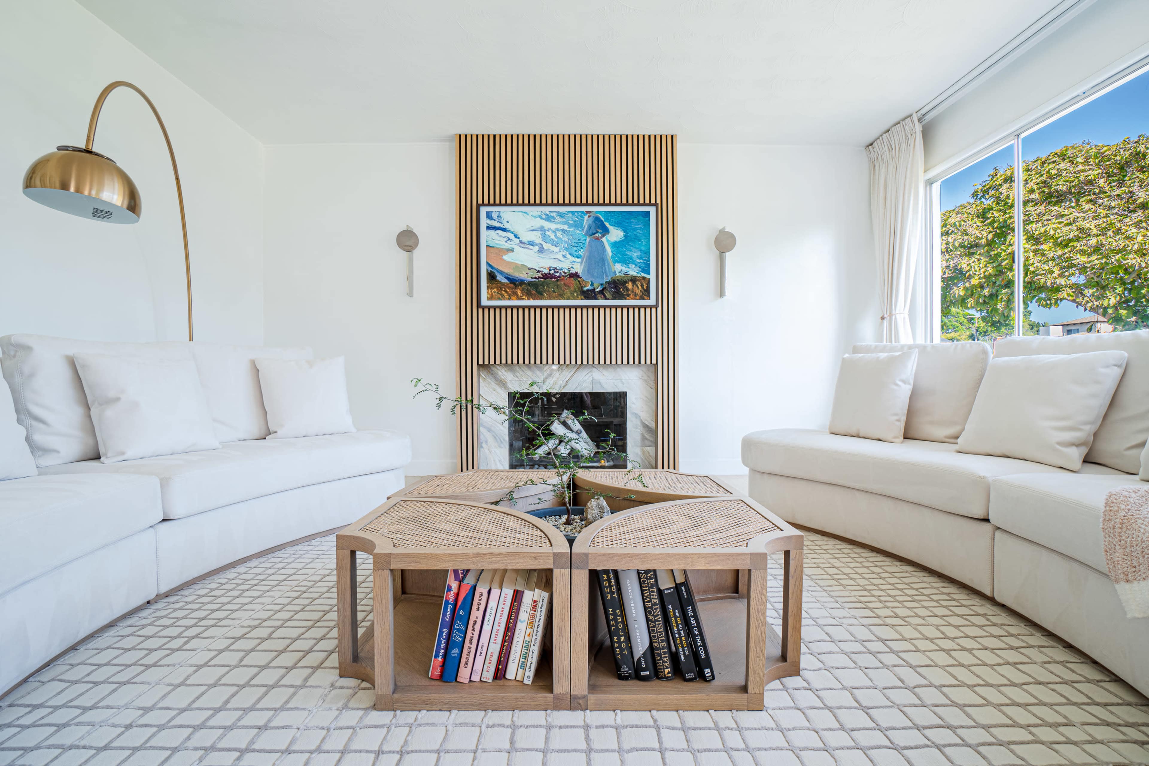 The image features a modern living room with two curved sofas, a central coffee table stacked with books, and a painting above a fireplace.
