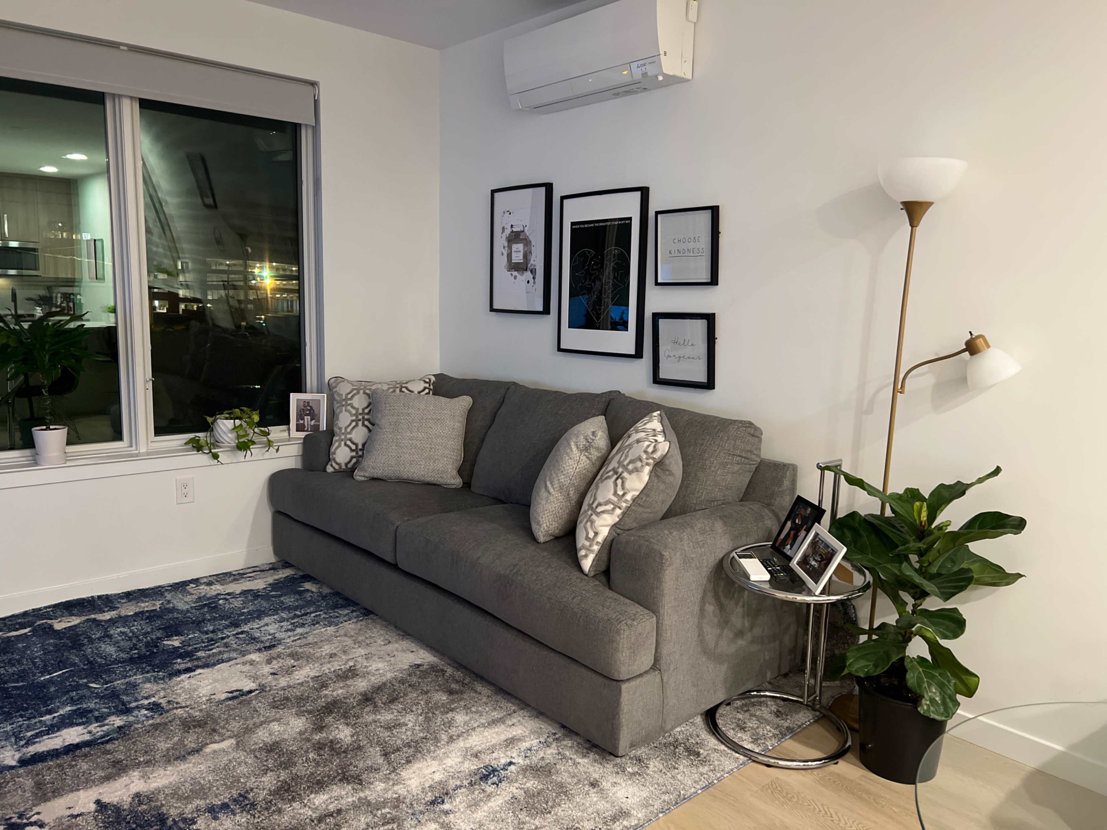 A gray sectional sofa is positioned against a white wall, surrounded by framed artwork and a tall plant, with a lamp and decorative items on a side table.