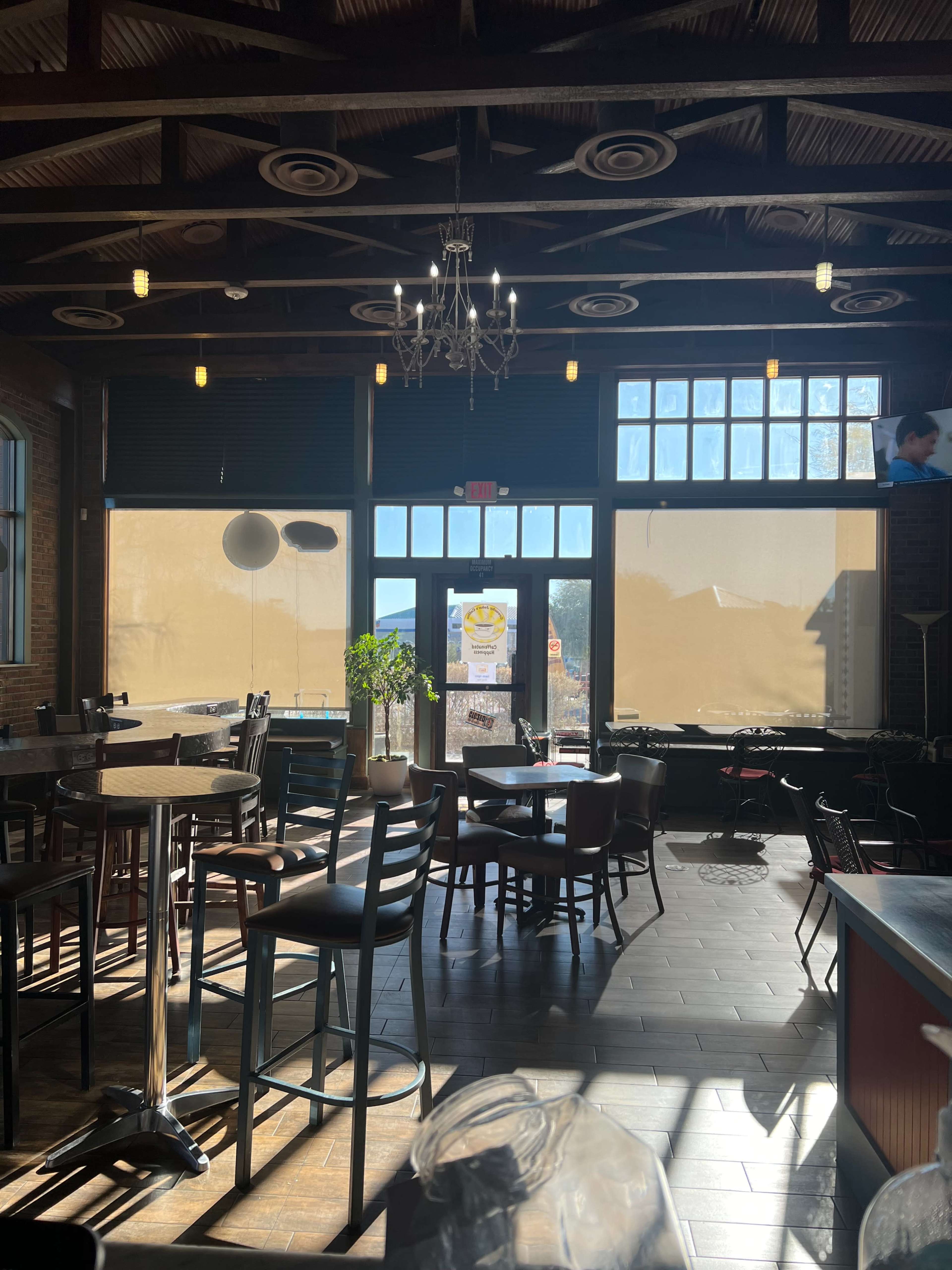 The interior of a restaurant with wooden furniture, large windows covered partially with shades, and hanging light fixtures.