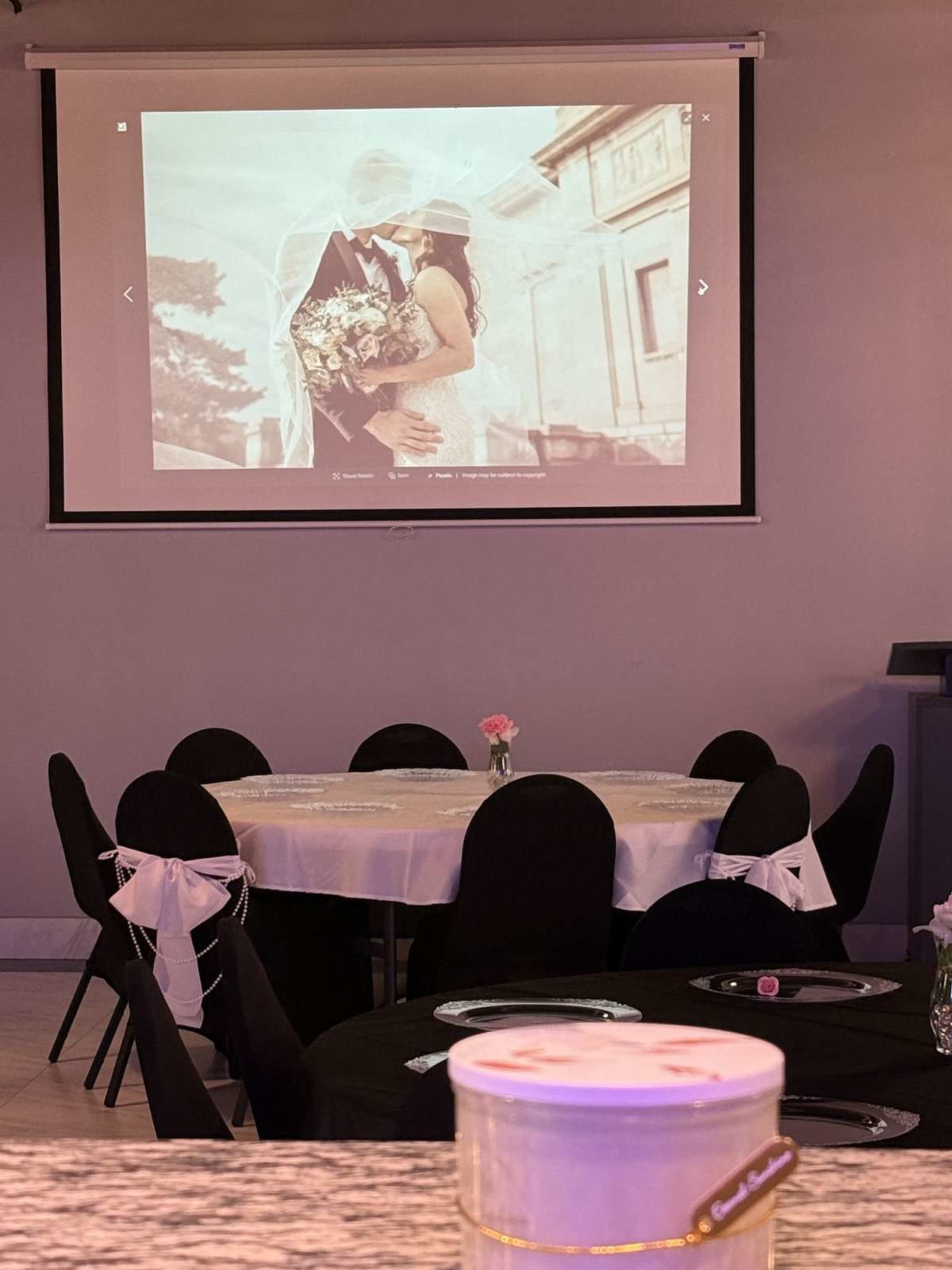 A wedding reception setup is shown, featuring round tables with black tablecloths and white decor, while a large screen displays a couple embracing.