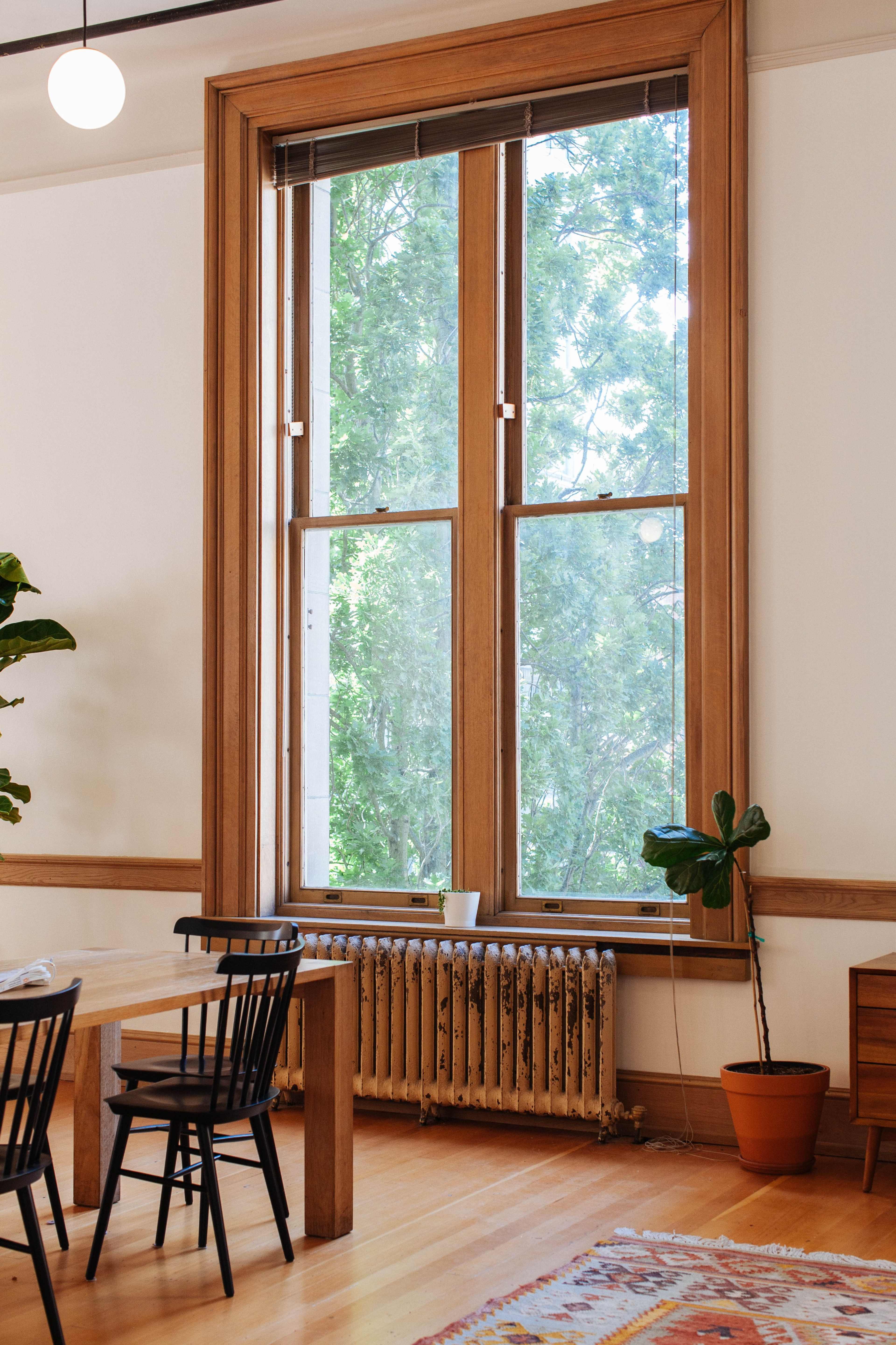 A large window with wooden frames and a view of greenery overlooks a dining area featuring a table and black chairs.