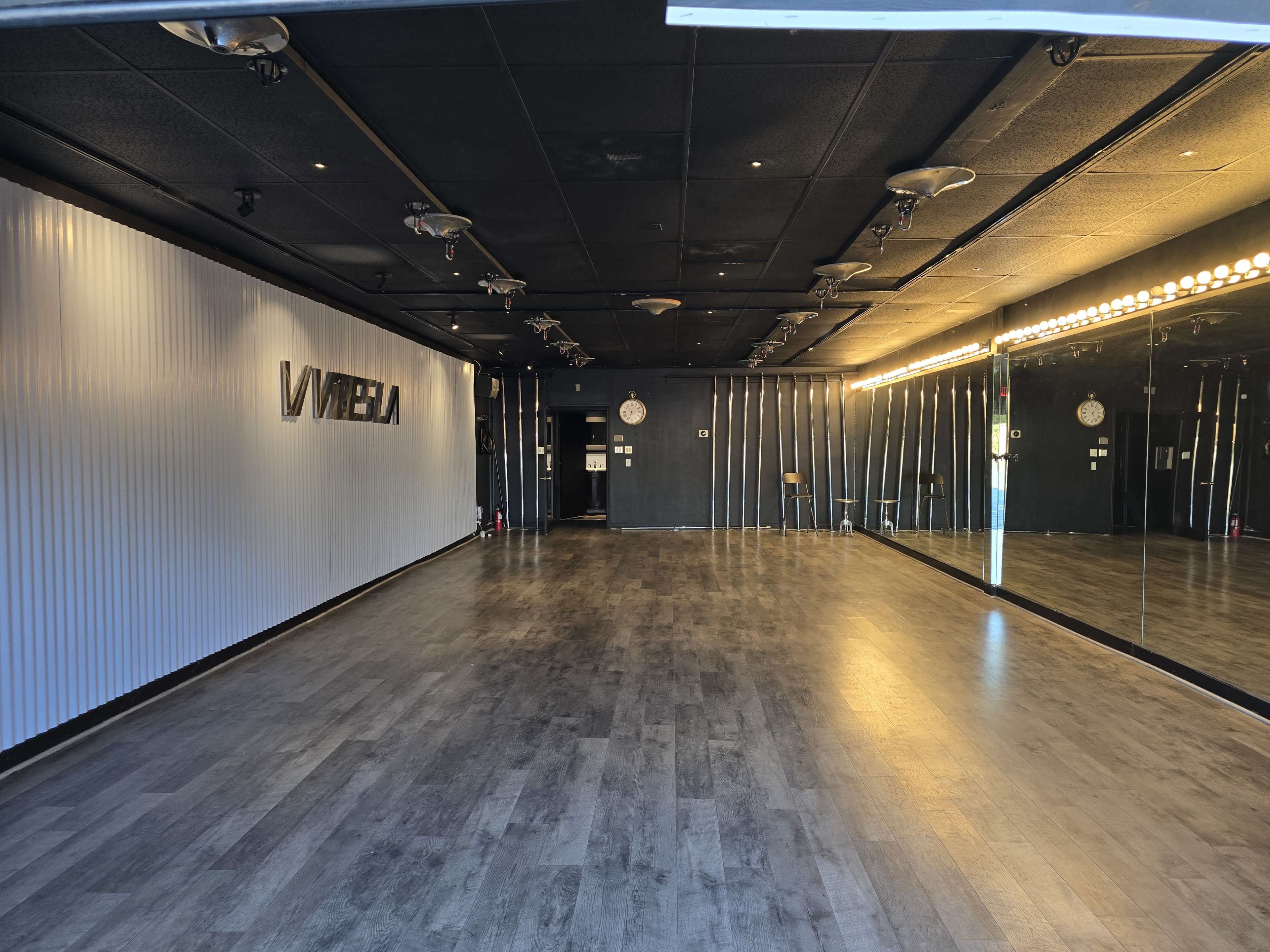 The image shows an empty dance studio with a mirrored wall, wooden flooring, and a lit vanity mirror on one side.