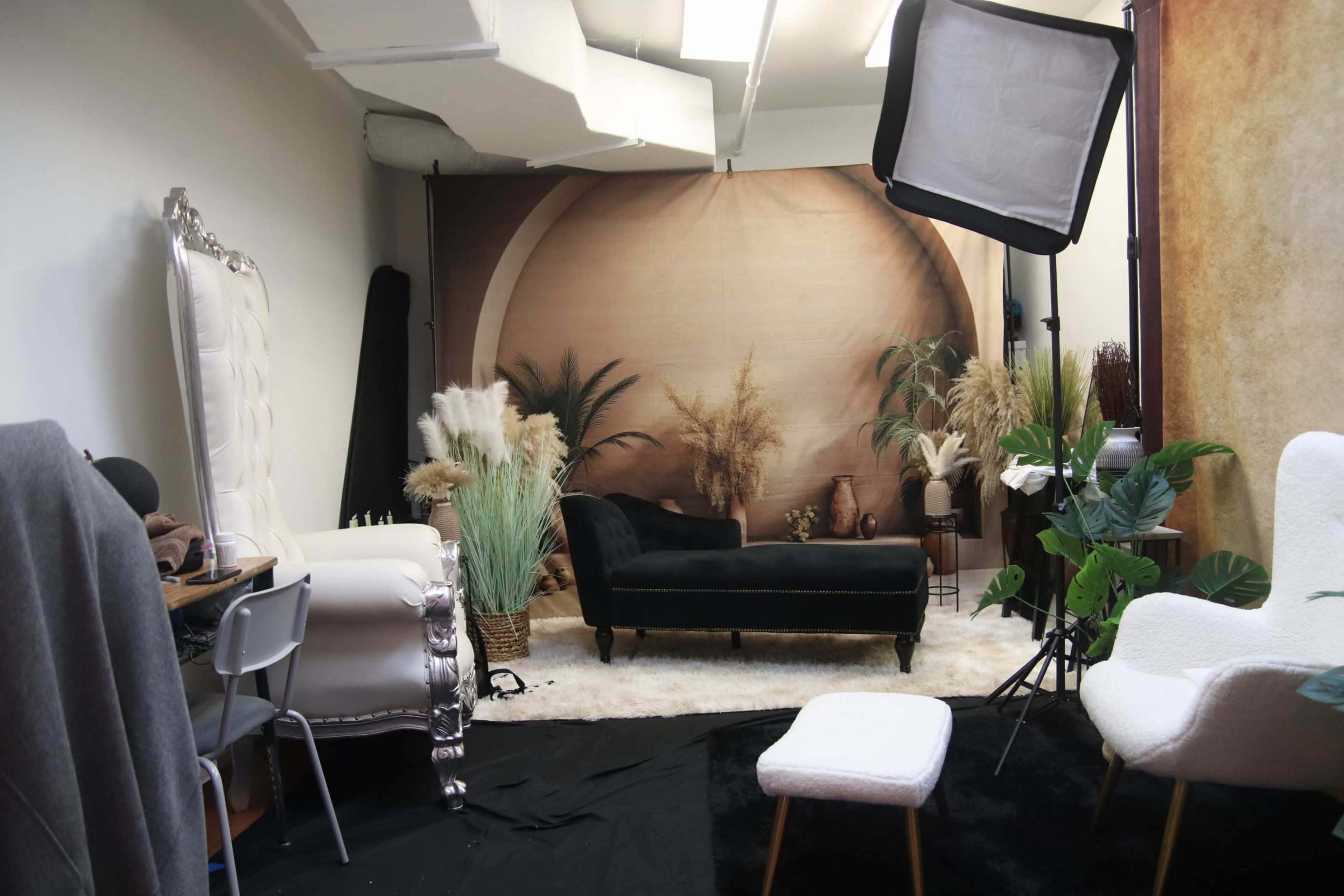 The image shows a styled photography studio featuring a black velvet chaise lounge, various decorative plants, and a backdrop with a neutral tone, complemented by soft furnishings and lighting equipment.