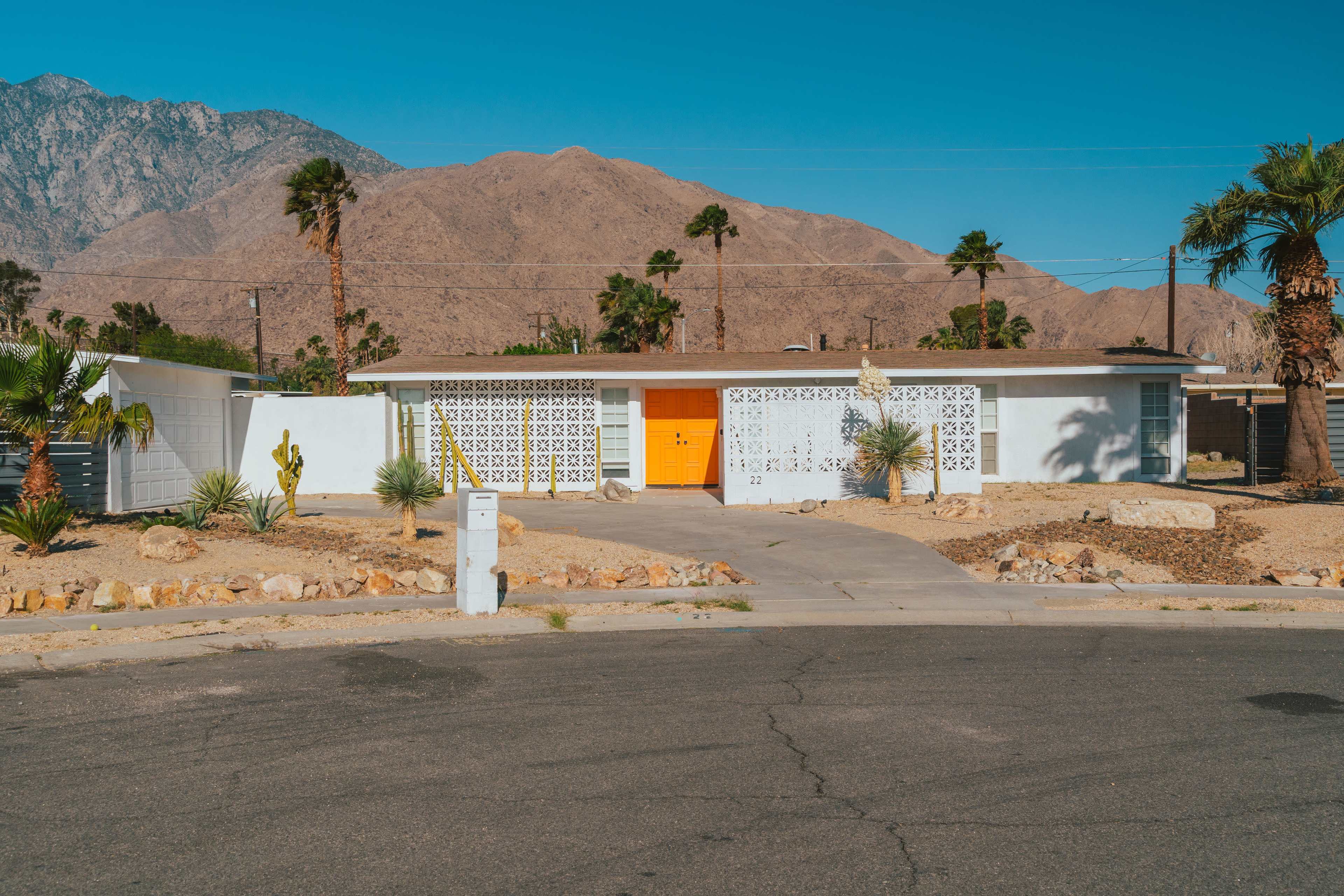 A single-story white house with an orange door is situated against a backdrop of mountains, surrounded by desert landscaping and palm trees.