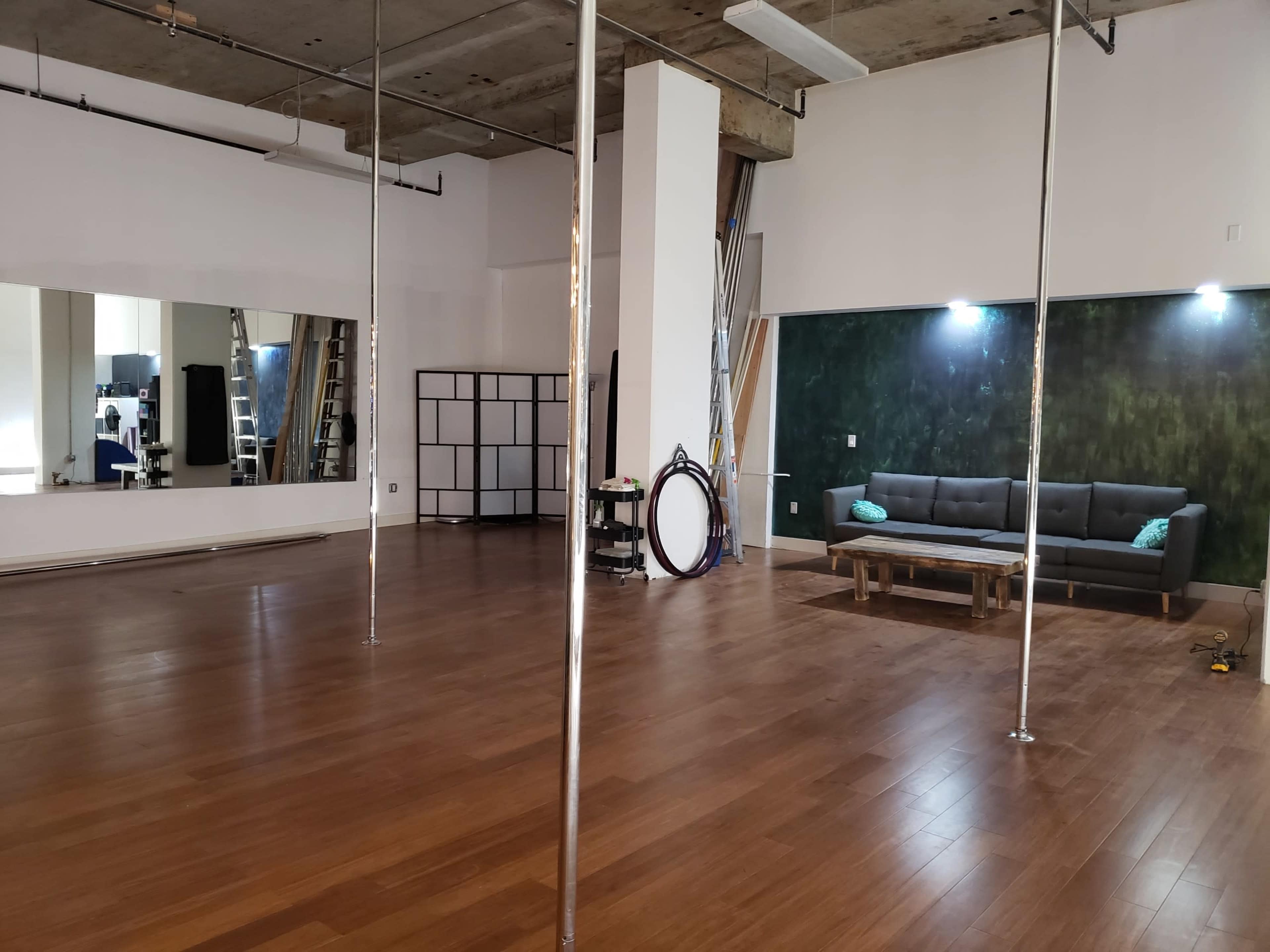 A spacious studio with wooden flooring, featuring several pole dance apparatuses, a mirror on one wall, and a sofa set against a green accent wall.