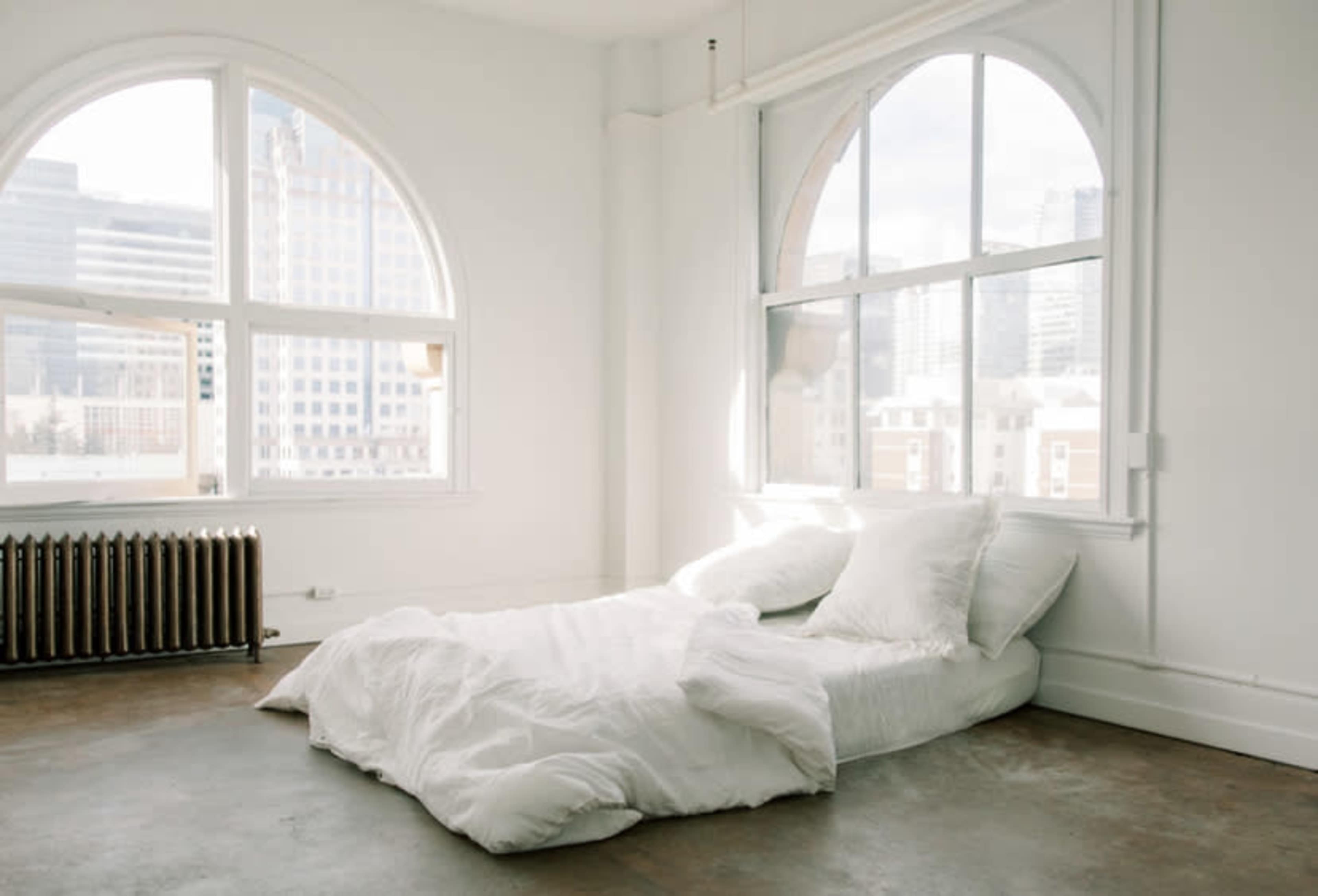 A minimalist bedroom features a unmade bed with white bedding in front of large arched windows overlooking a city skyline.