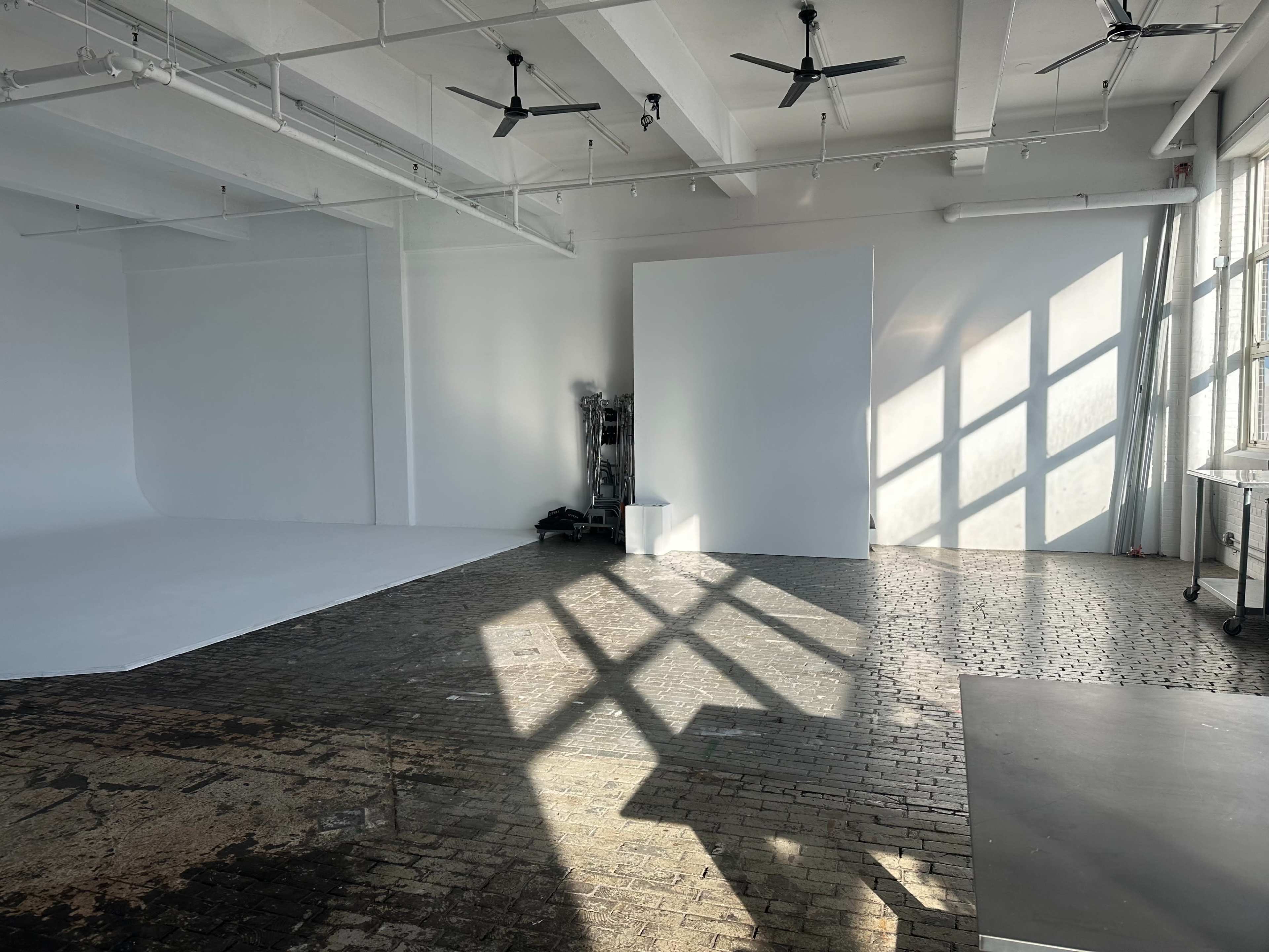 A spacious, well-lit photography studio with a white backdrop, large windows casting shadows on the floor, and industrial ceiling fans.