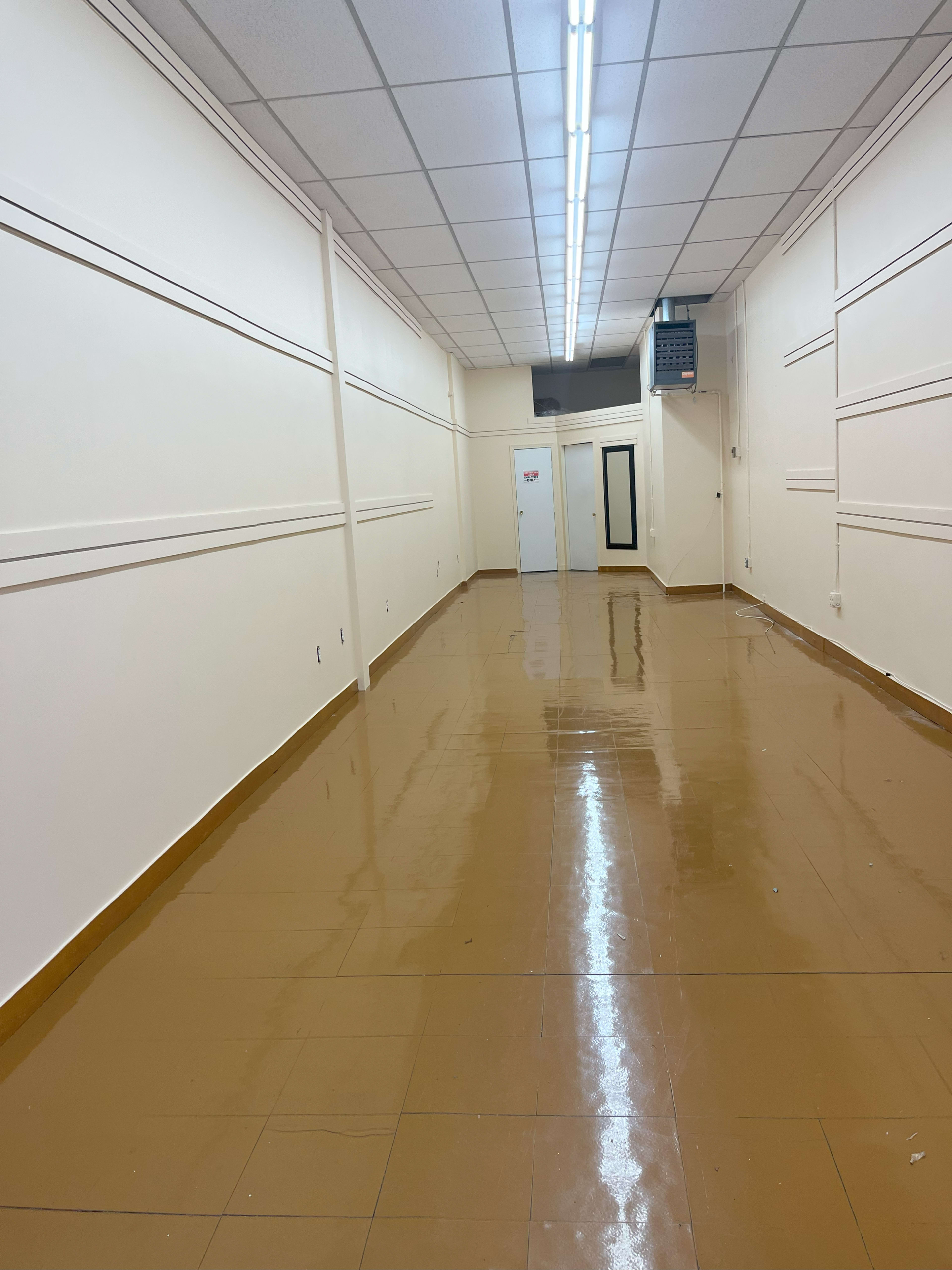 A long, empty hallway with a glossy, tiled floor and plain walls, leading to a door at the far end.
