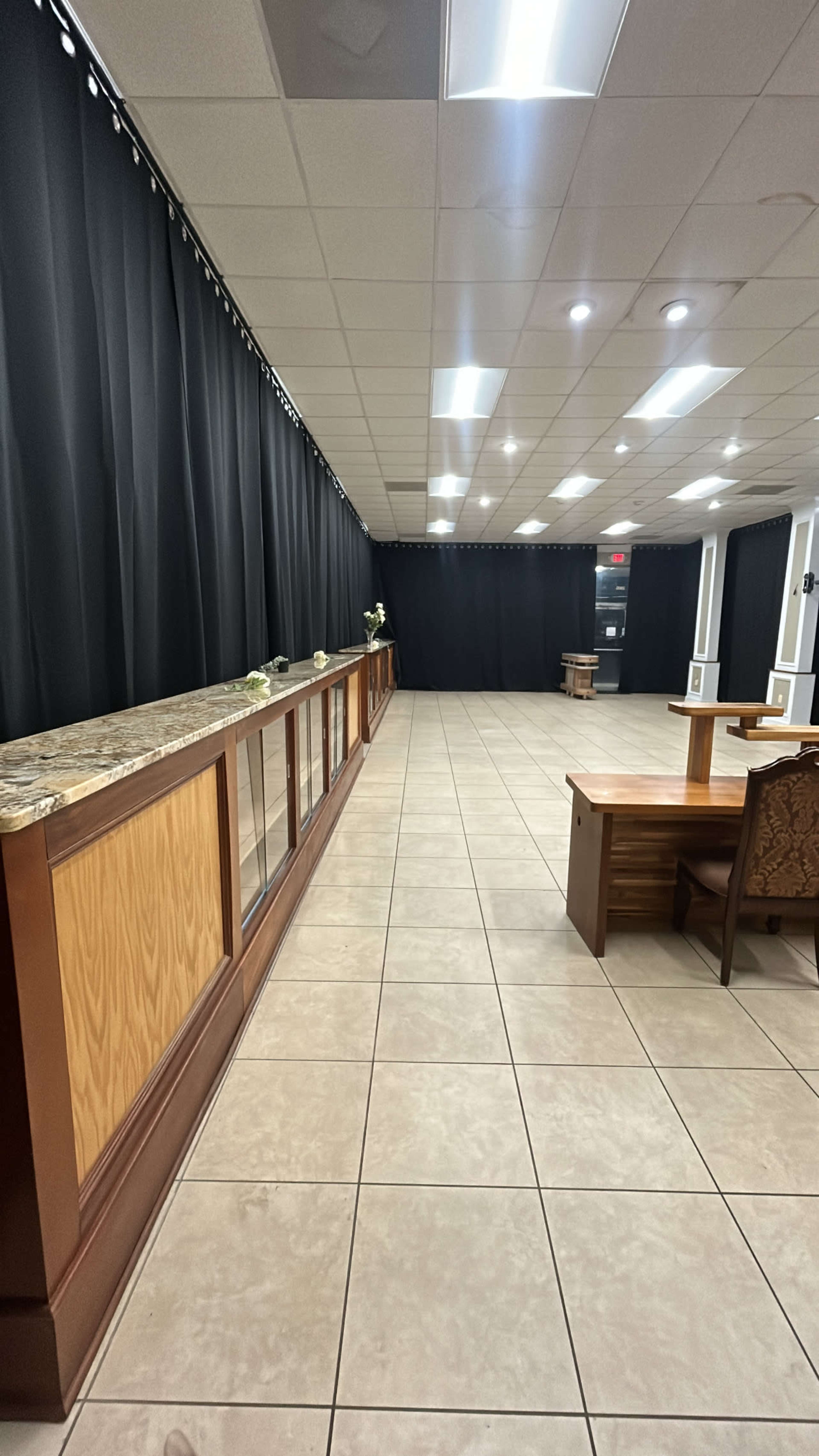 A spacious room with tiled flooring, a long wooden counter against one wall, and a table in the center, surrounded by black curtains.
