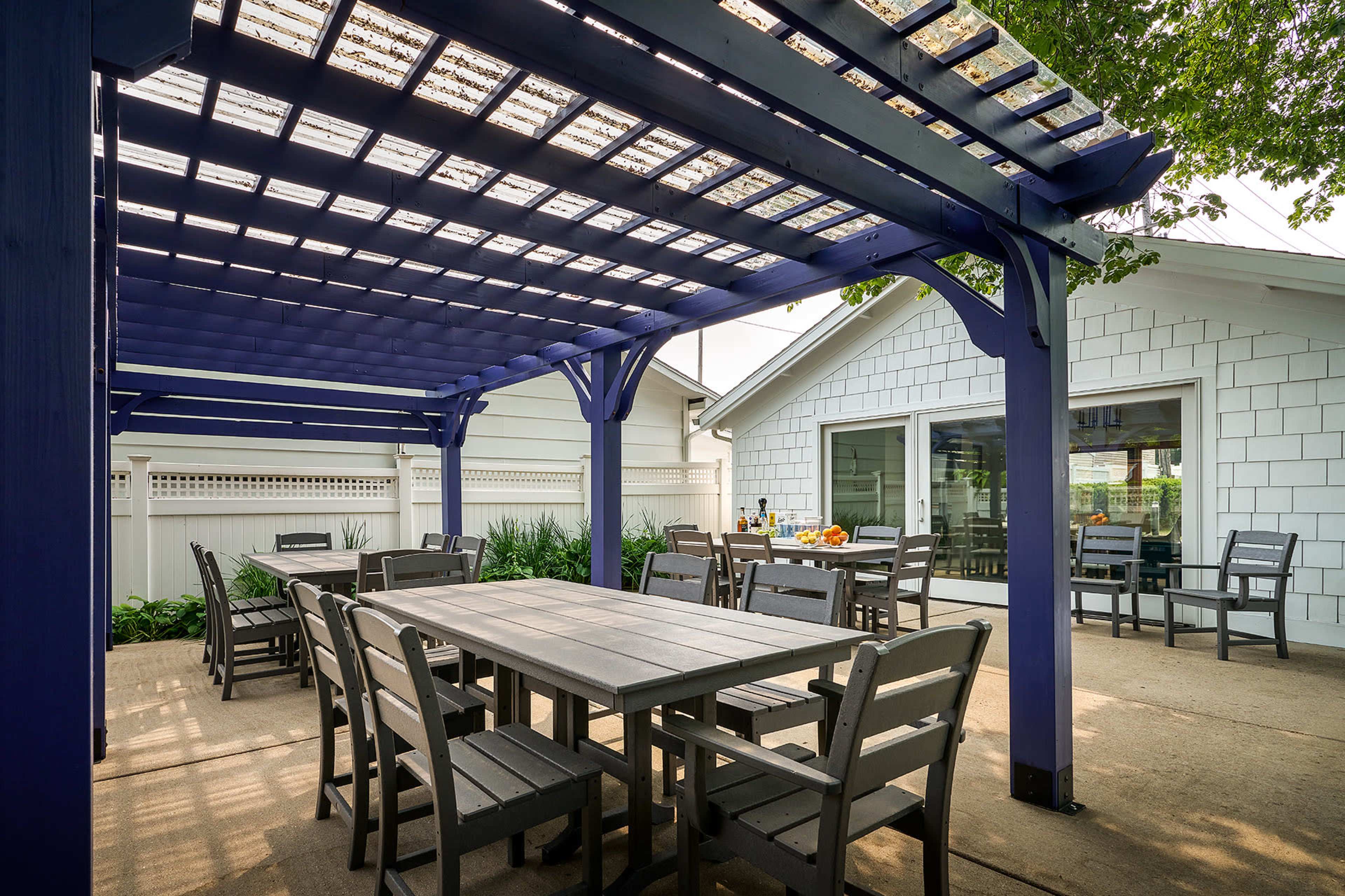 A spacious outdoor dining area with a purple pergola, wooden tables and chairs, and a house in the background.