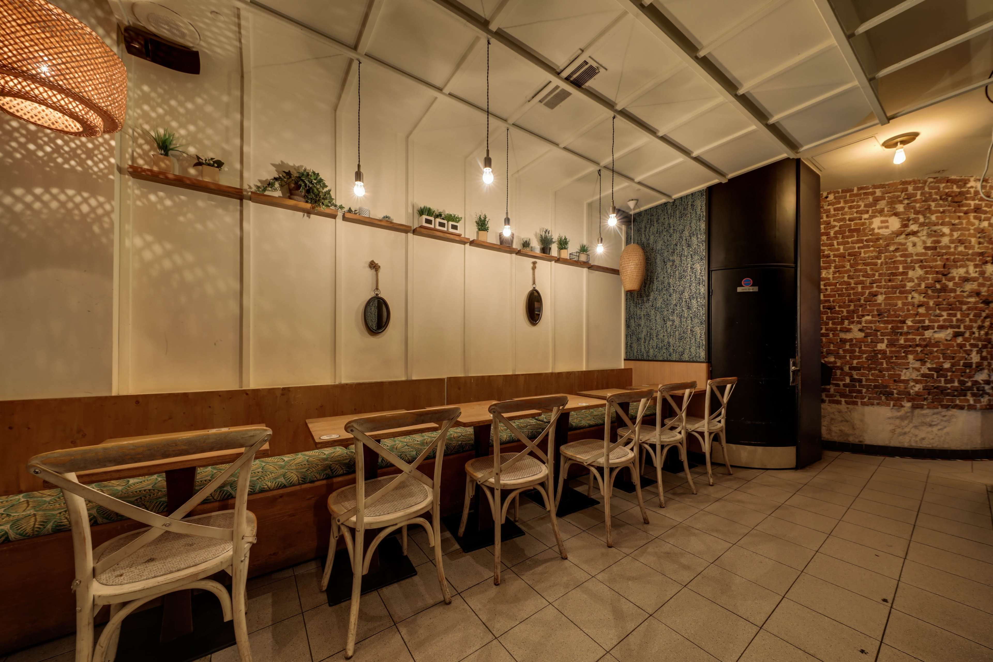 The image shows a modern café interior with a long wooden table surrounded by white chairs, set against a wall featuring decorative plants and a brick accent wall.