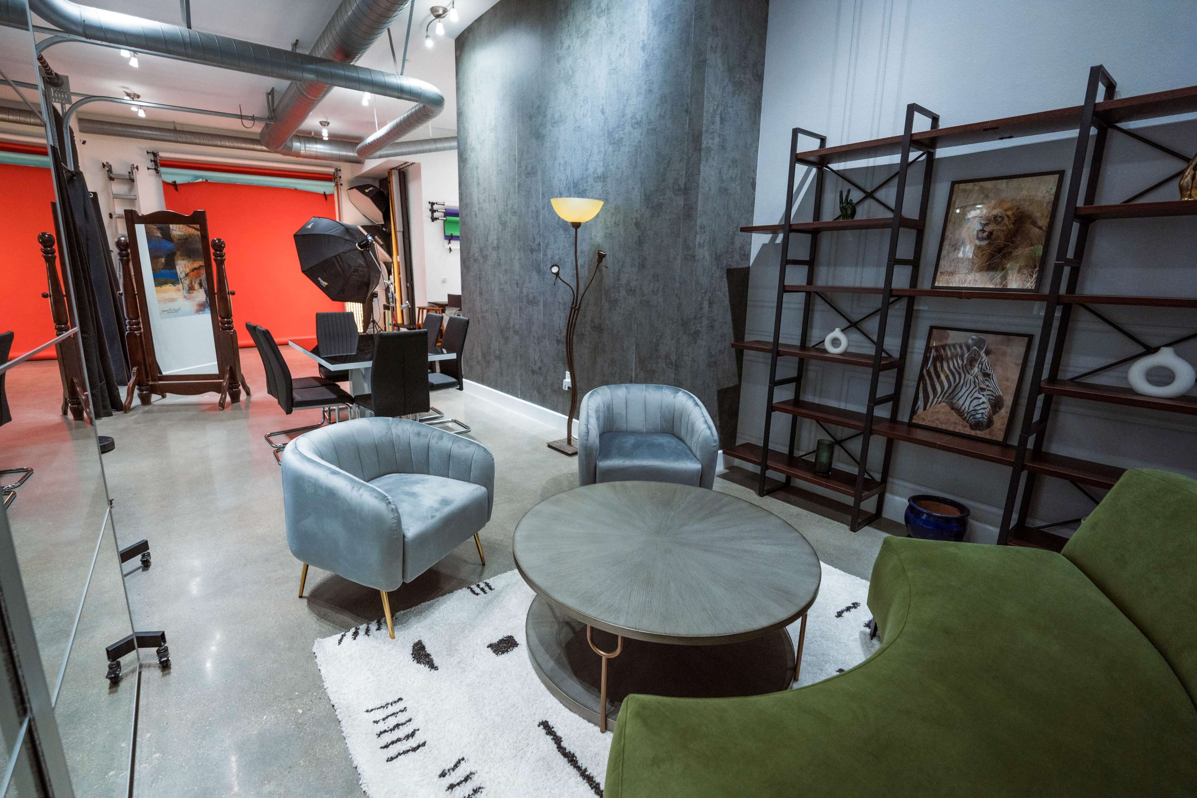 The image shows a modern studio space featuring a seating area with two armchairs and a coffee table, surrounded by a dining area and photography equipment against a backdrop of orange and gray walls.