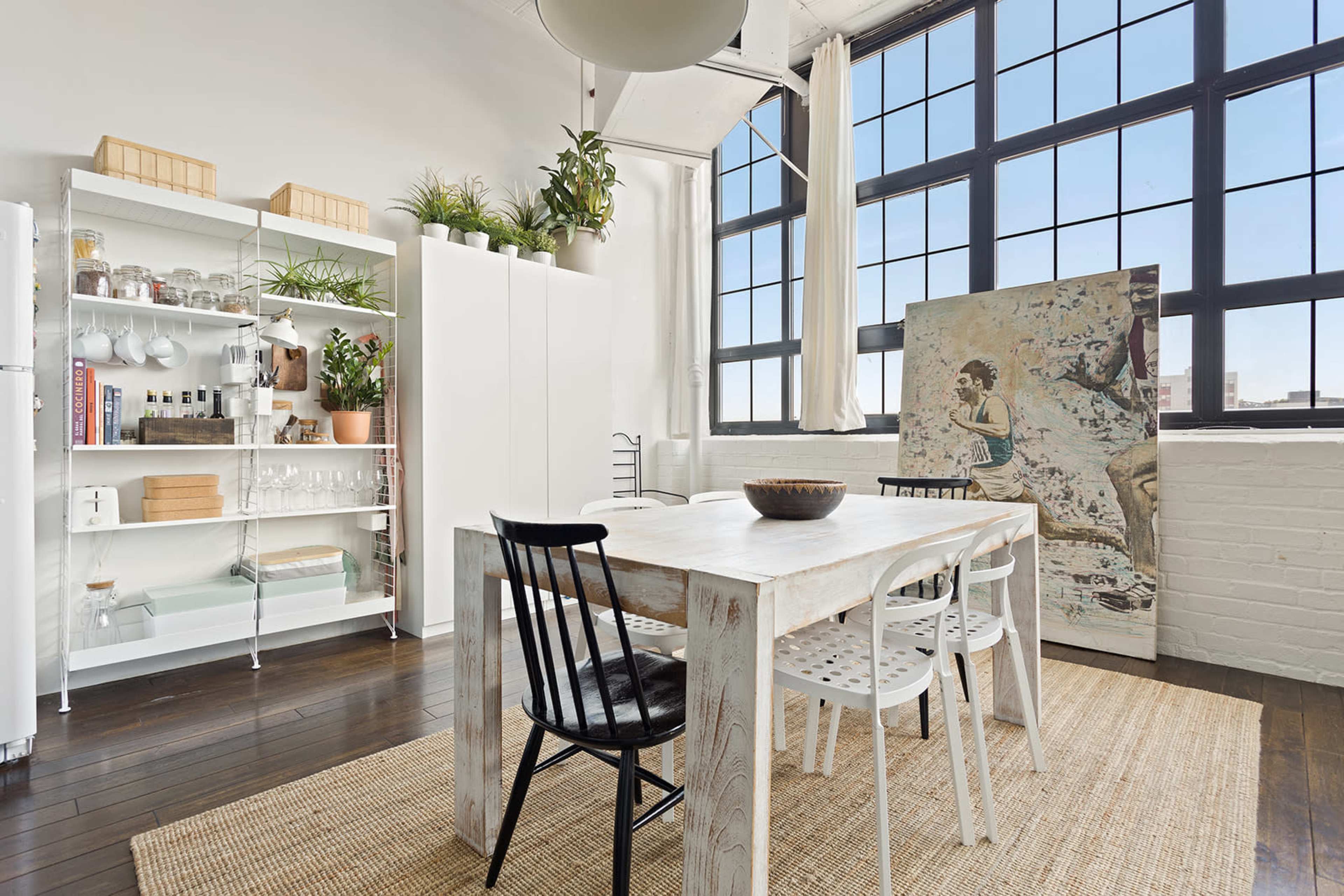 A dining area features a white wooden table surrounded by black and white chairs, with large windows and a contemporary art piece nearby.