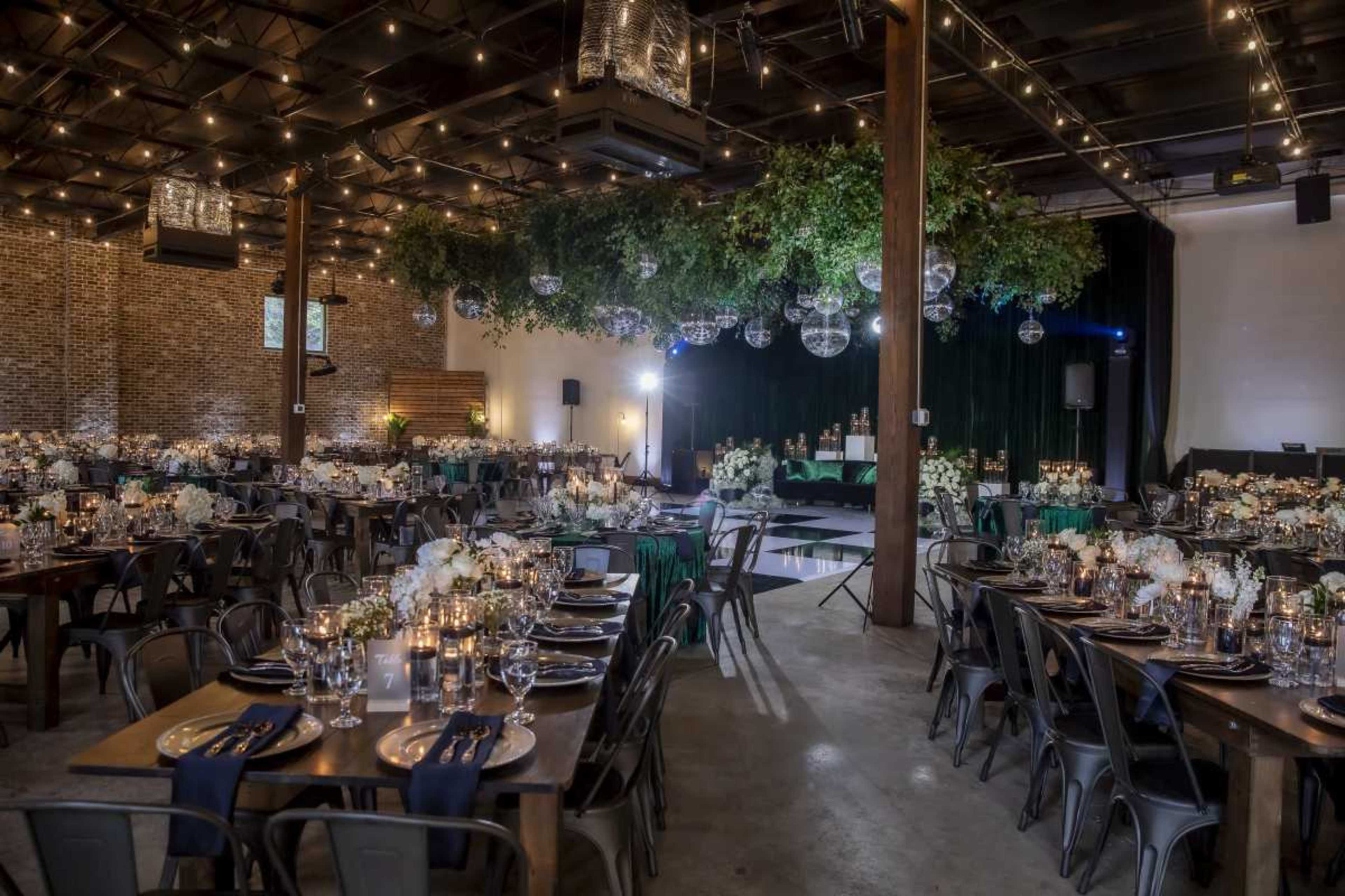 The image shows a spacious event venue set up for a formal gathering, featuring multiple tables adorned with flowers and candles, and a stage area decorated with greenery and lighting fixtures.