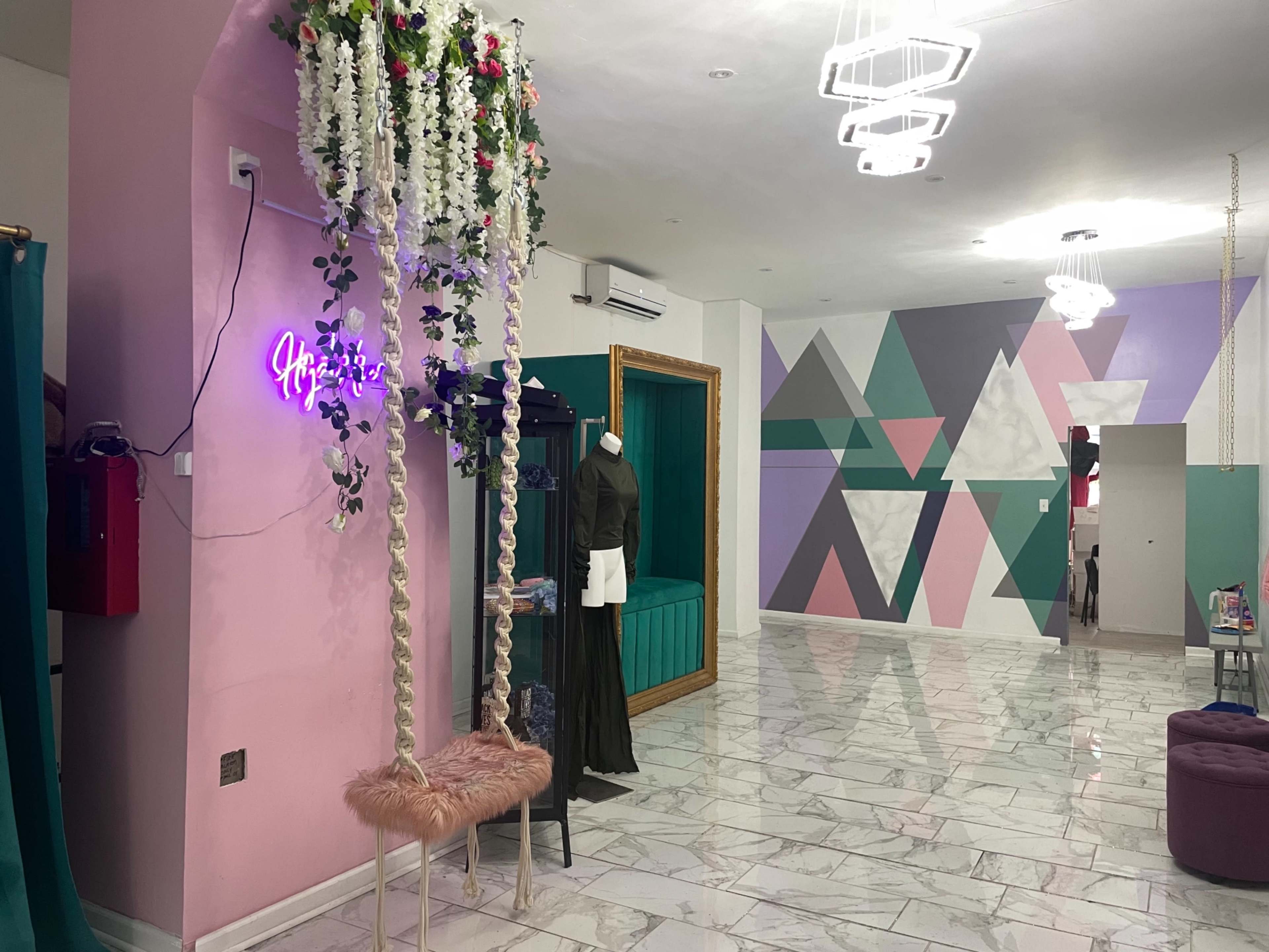 A modern boutique interior with a pink accent wall, a floral swing, a mannequin displaying clothing, and a geometric mural painted on the far wall.