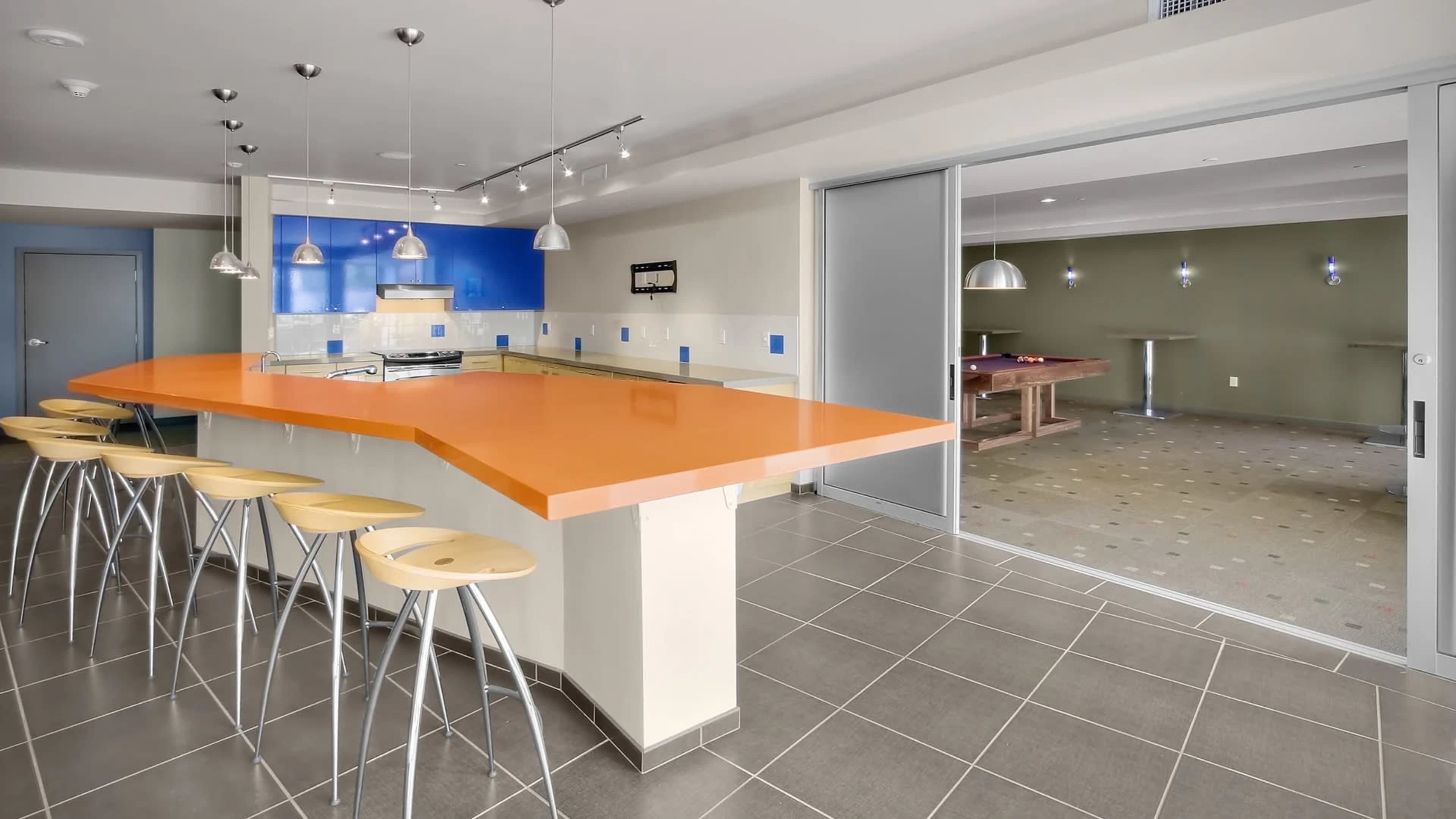 A modern kitchen features an orange countertop with bar stools and a view of a separate room containing a pool table.