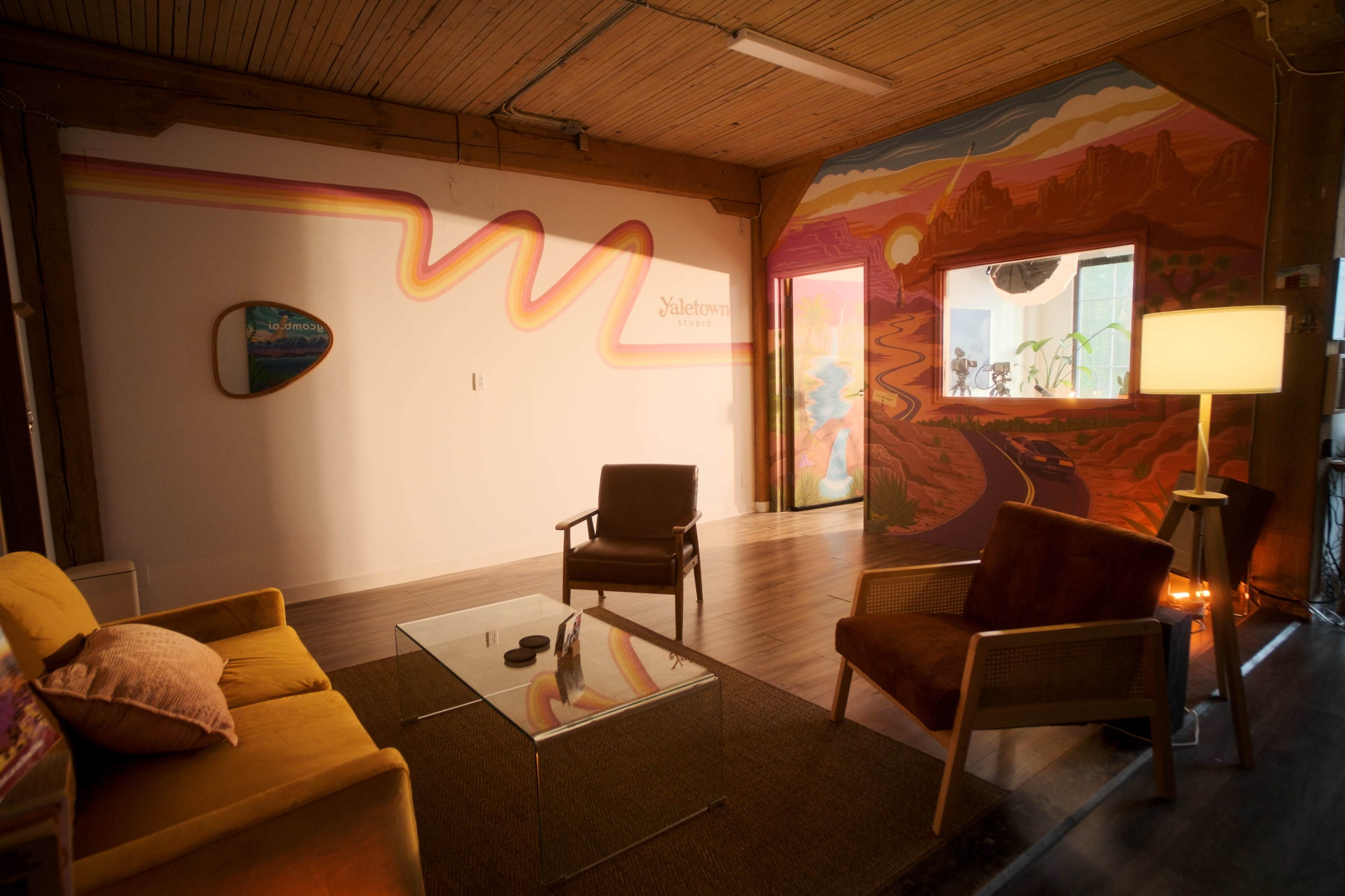 The image shows a brightly colored workspace with a cozy seating area featuring a yellow couch, brown armchairs, and a glass table, alongside a vibrant mural depicting a scenic landscape.