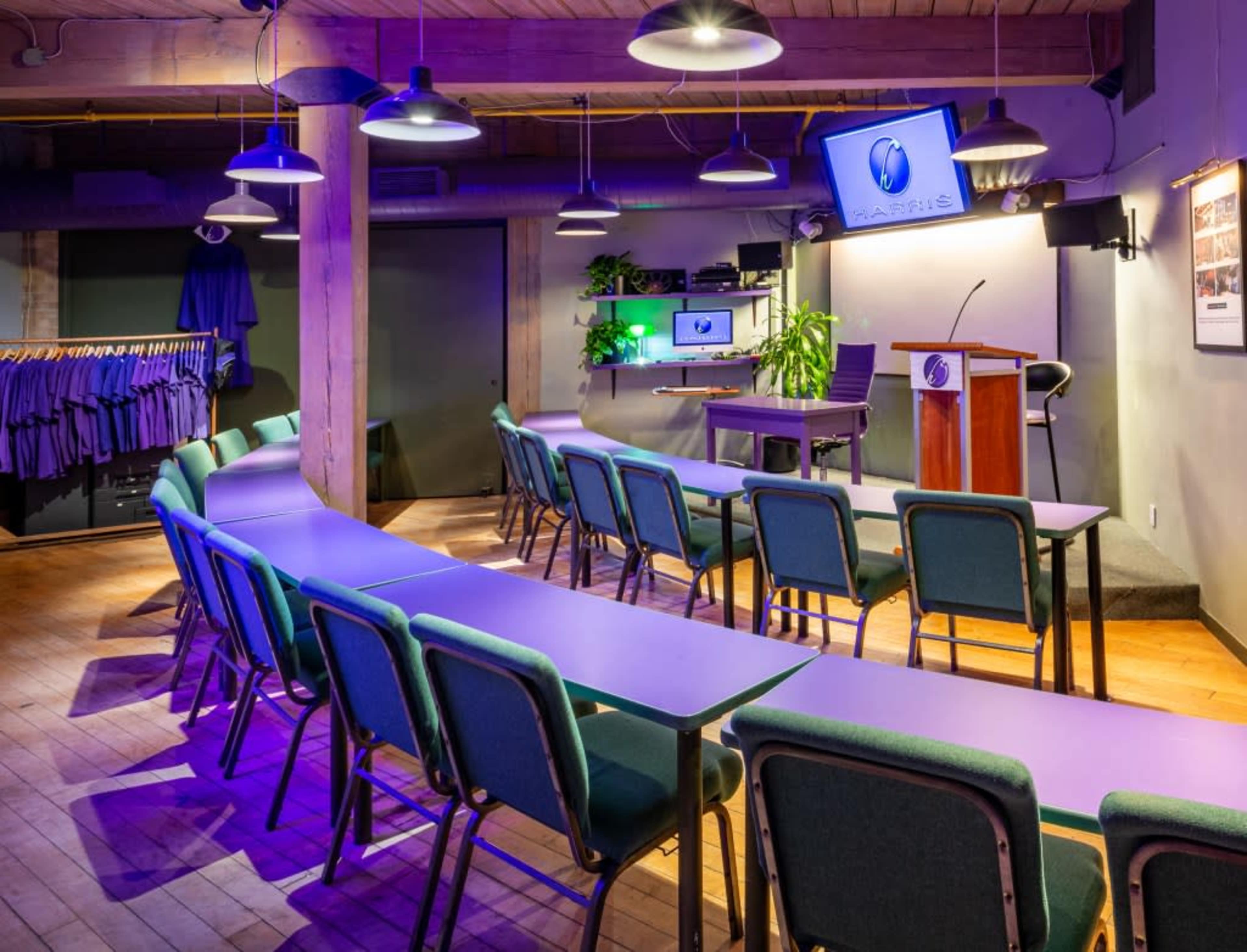 A classroom or meeting space with rows of tables and chairs arranged facing a podium and screens, surrounded by plants and hanging lights.