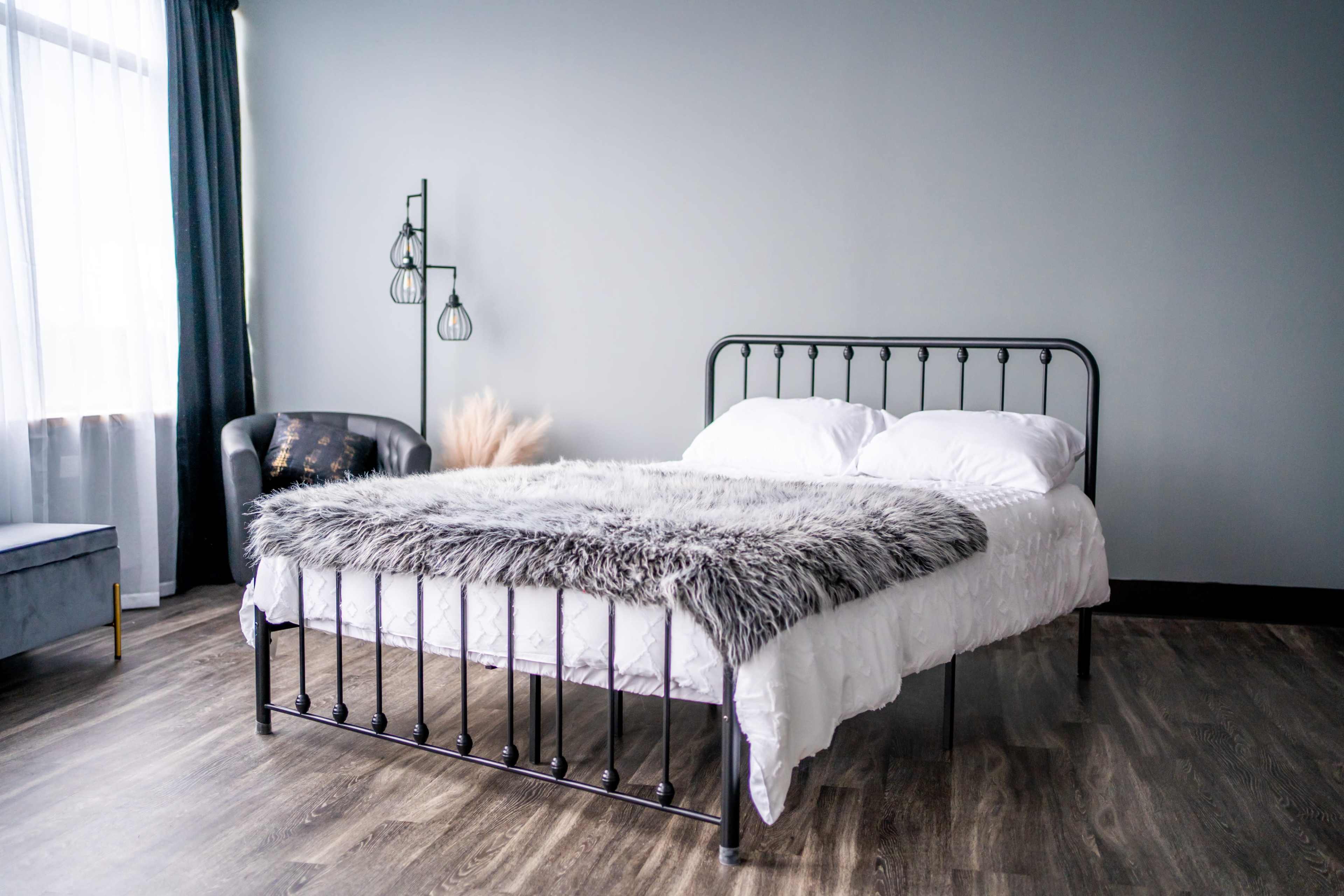 A simple bedroom features a metal bed with white bedding and a gray fur throw, accompanied by a black armchair and large windows.