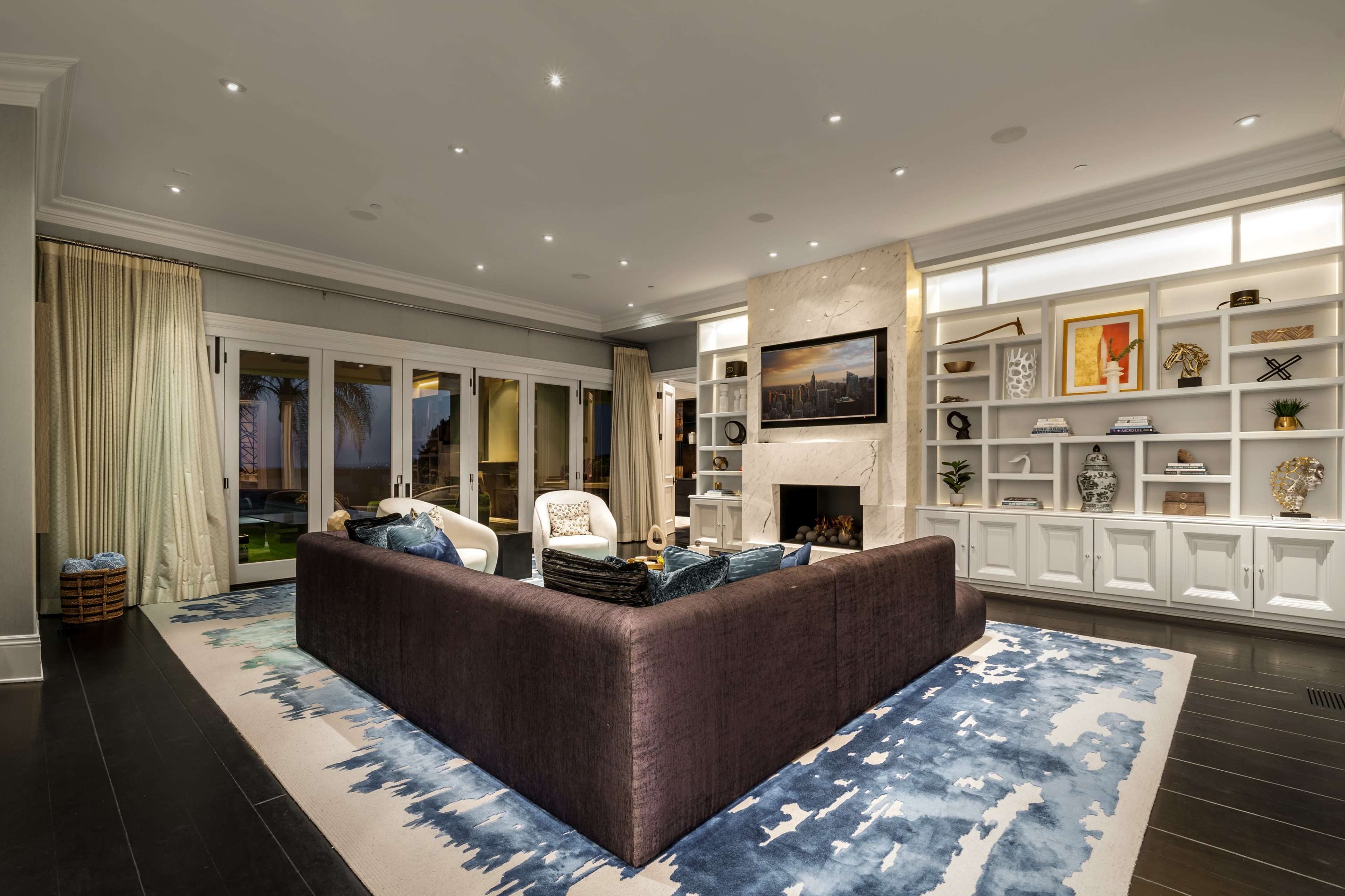A modern living room featuring a sectional sofa, a large area rug, built-in shelves with decorative items, and a fireplace, all illuminated by recessed lighting.