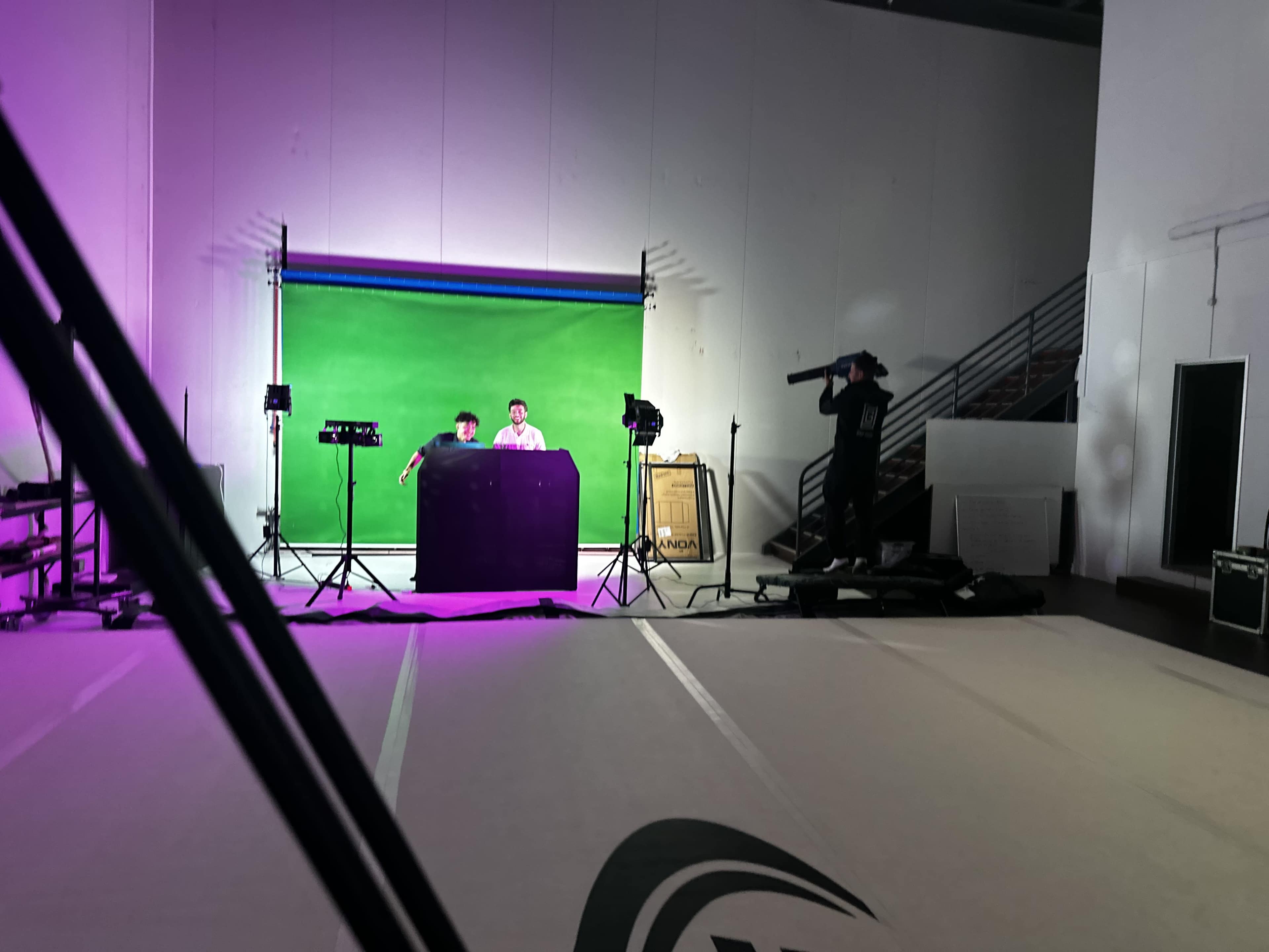A green screen setup is seen in a studio, where two individuals are positioned at a table while another person stands nearby with a camera.