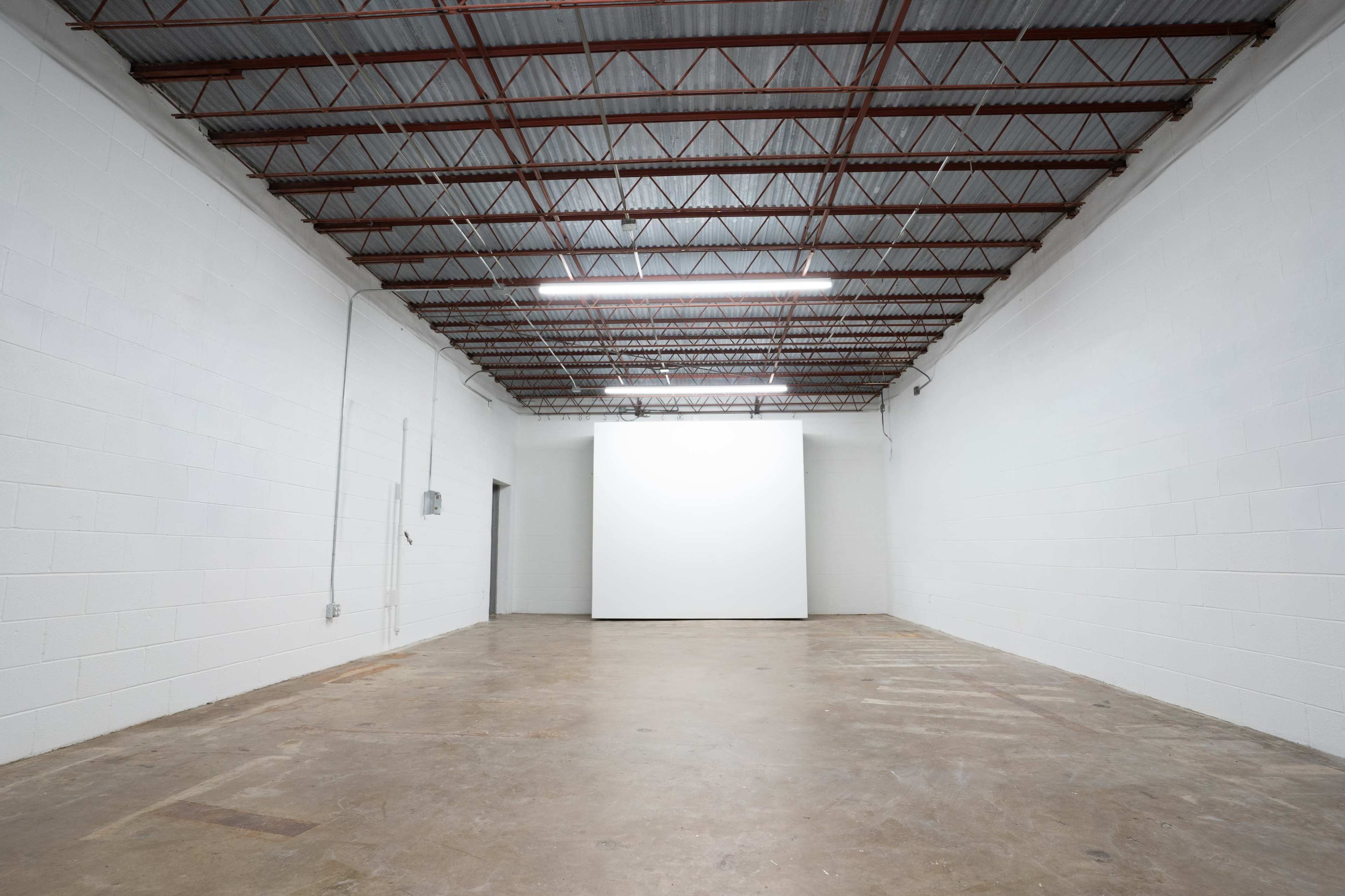 The image shows a large, empty white room with a concrete floor and a blank projection screen at one end.