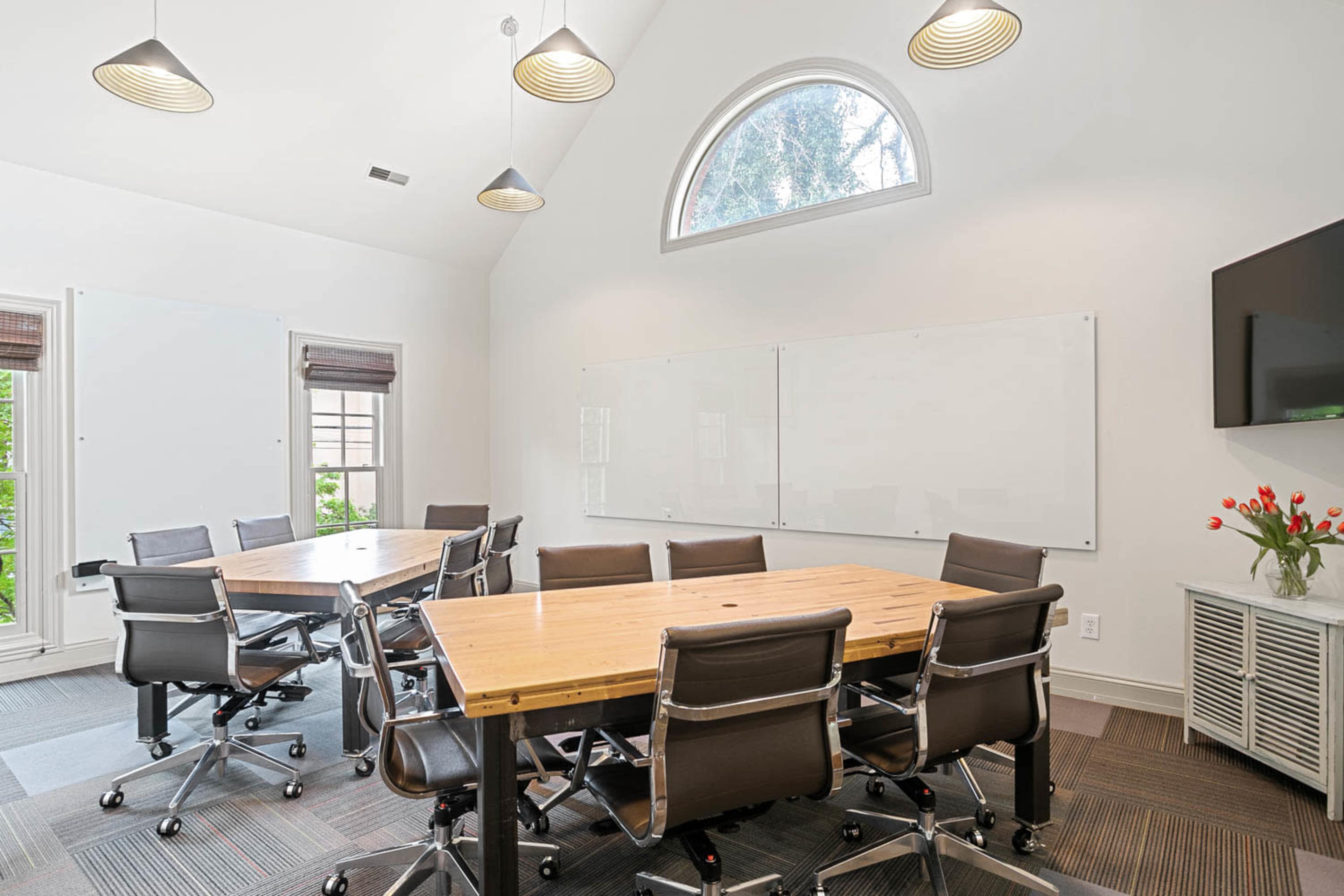 A modern conference room featuring a large wooden table surrounded by black rolling chairs, whiteboards on the walls, and a television mounted near a window.