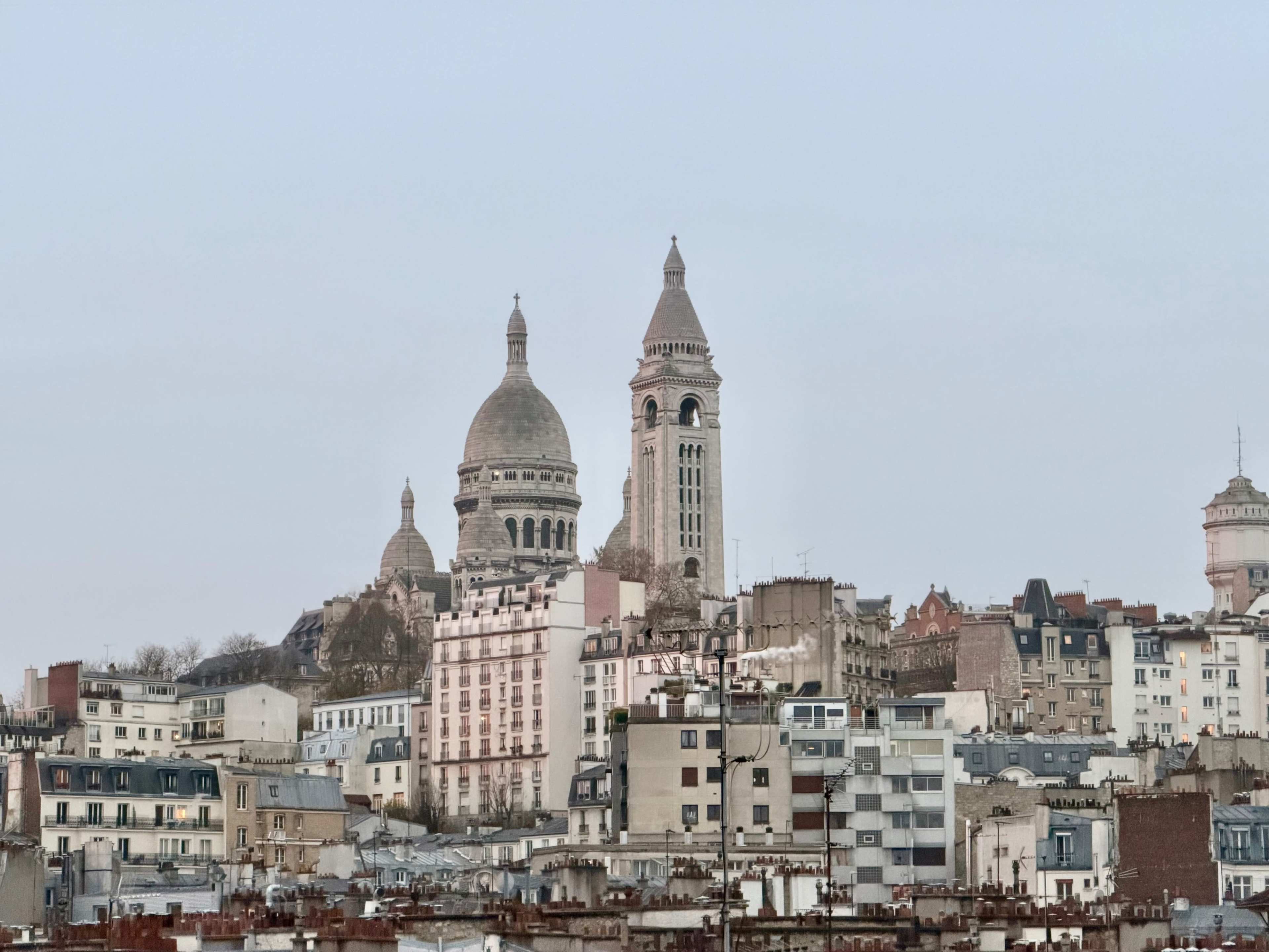 The image shows a view of the Sacré-Cœur Basilica on a hilltop, surrounded by various buildings in an urban landscape.