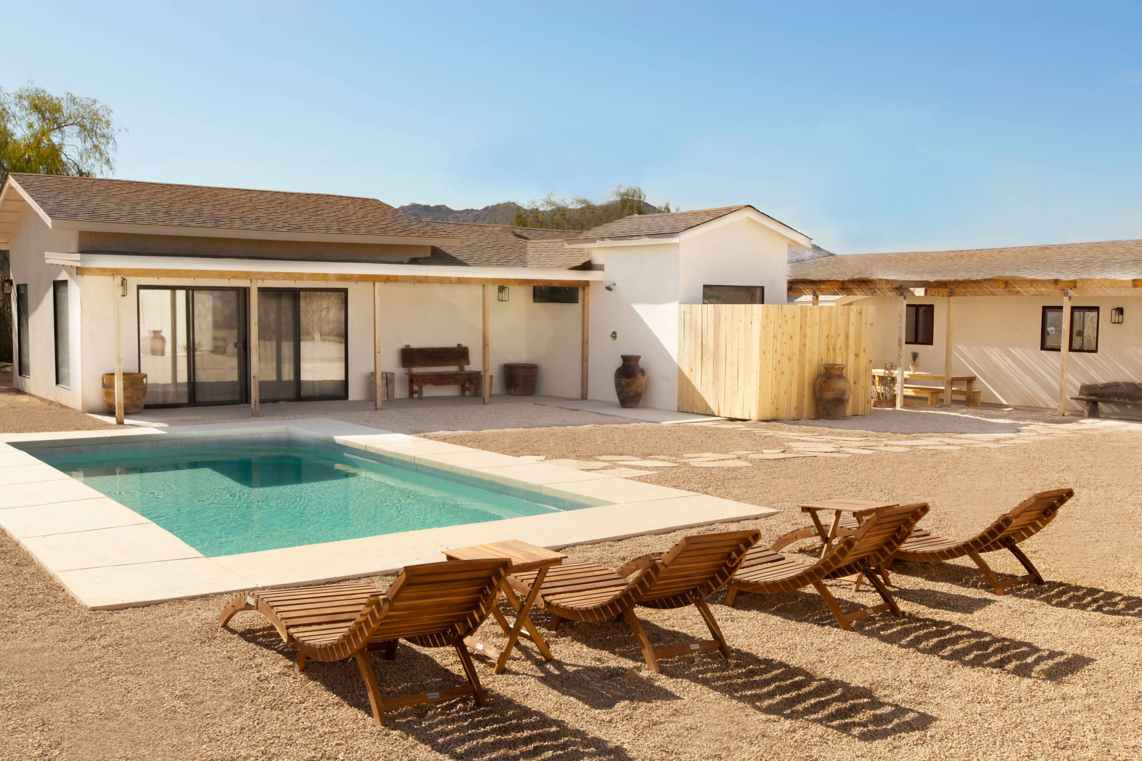 A modern white house with a pool and wooden lounge chairs is set in a gravel backyard surrounded by desert landscape.