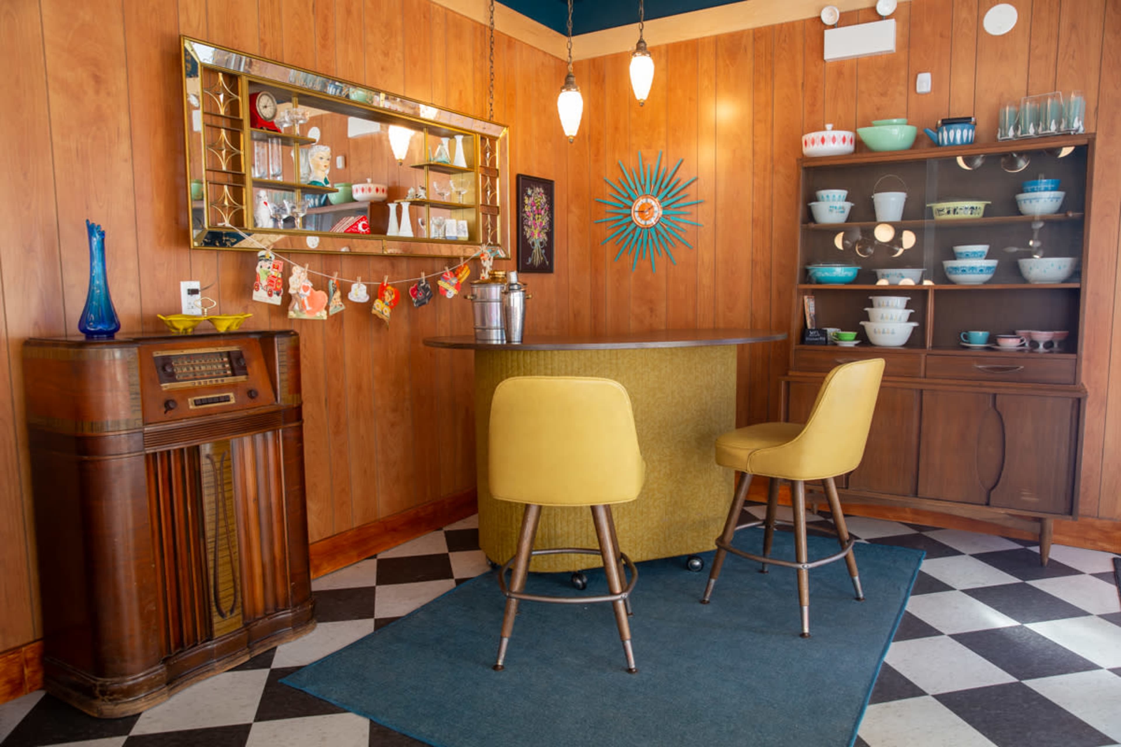 A vintage-style bar area with two yellow stools, a wooden console with a radio, a mirrored wall, and a display of colored dishware on a shelf.