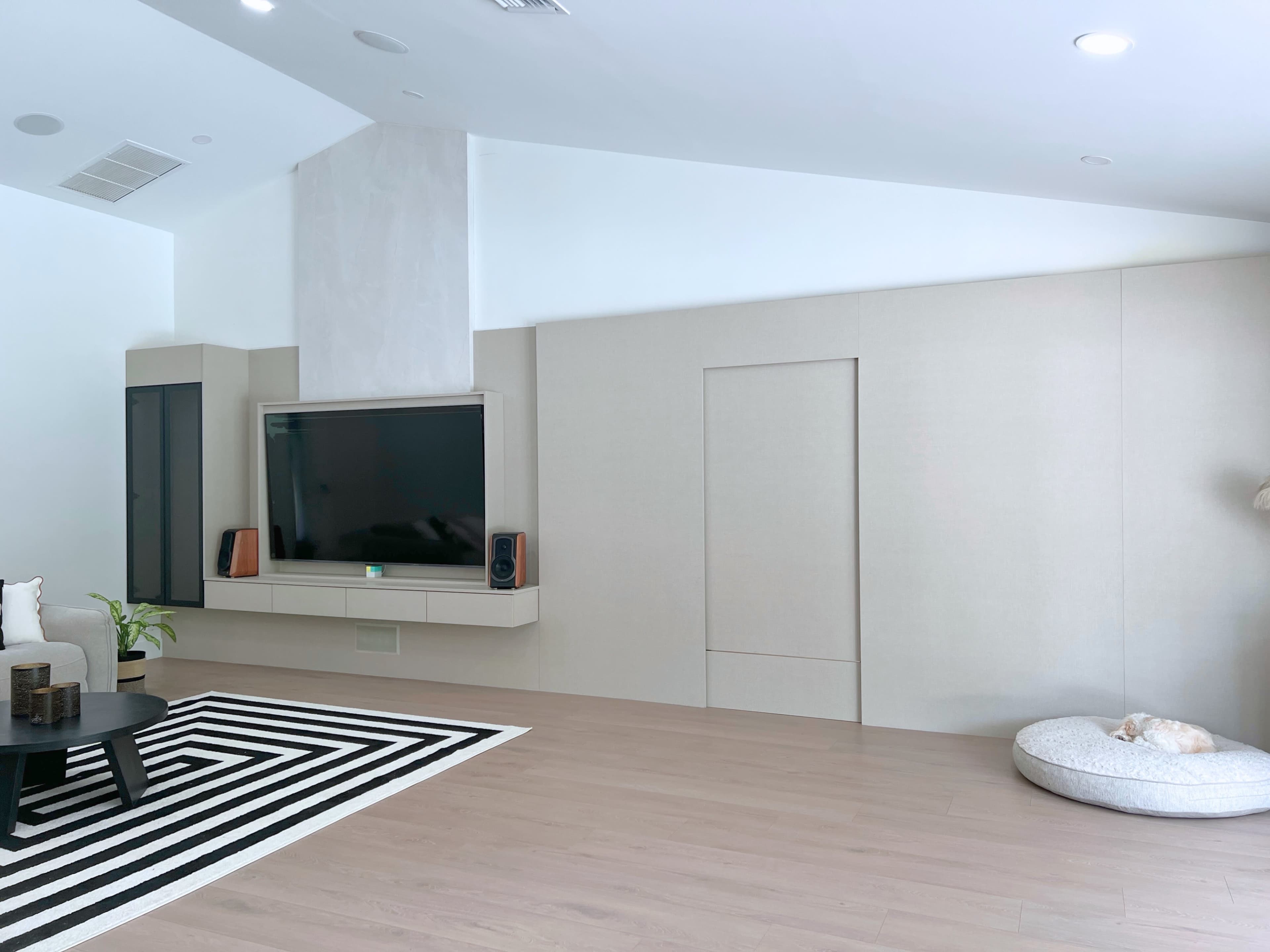 The image shows a modern living room with a minimalist design featuring a mounted television, a black and white striped rug, and a small pet bed in the corner.