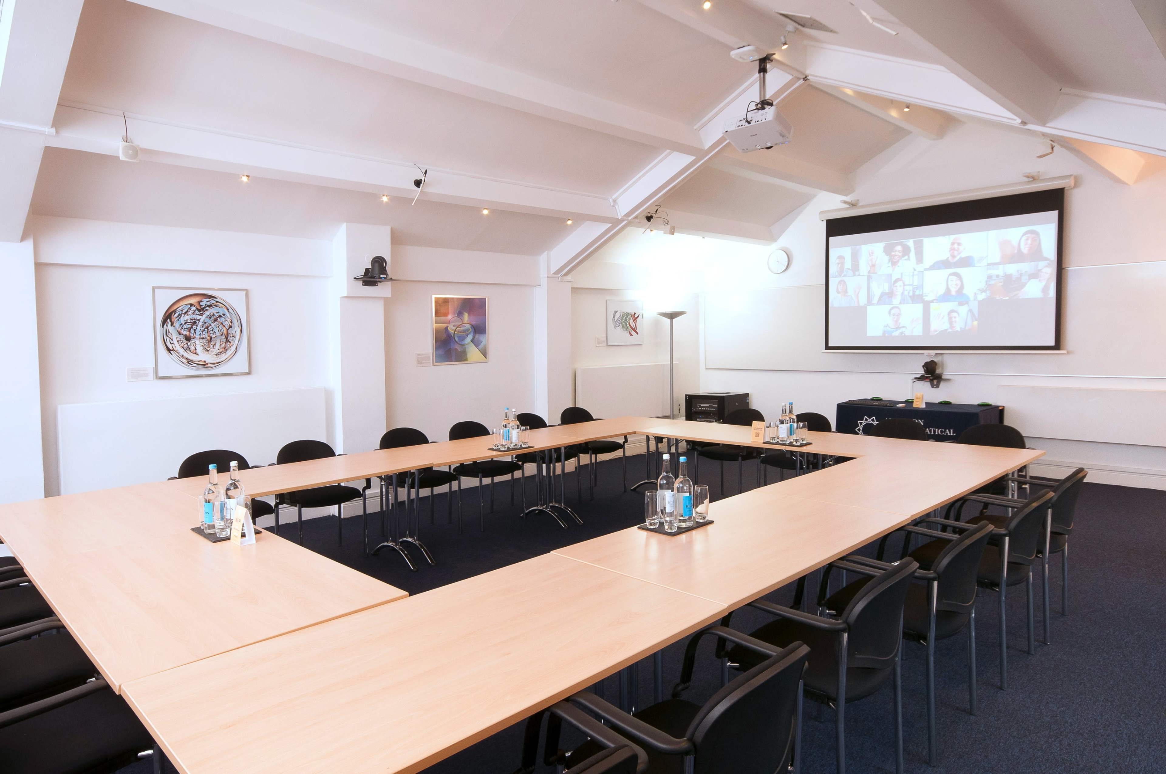 A modern conference room features a large table arranged in a U-shape, surrounded by black chairs, with a screen displaying a virtual meeting.