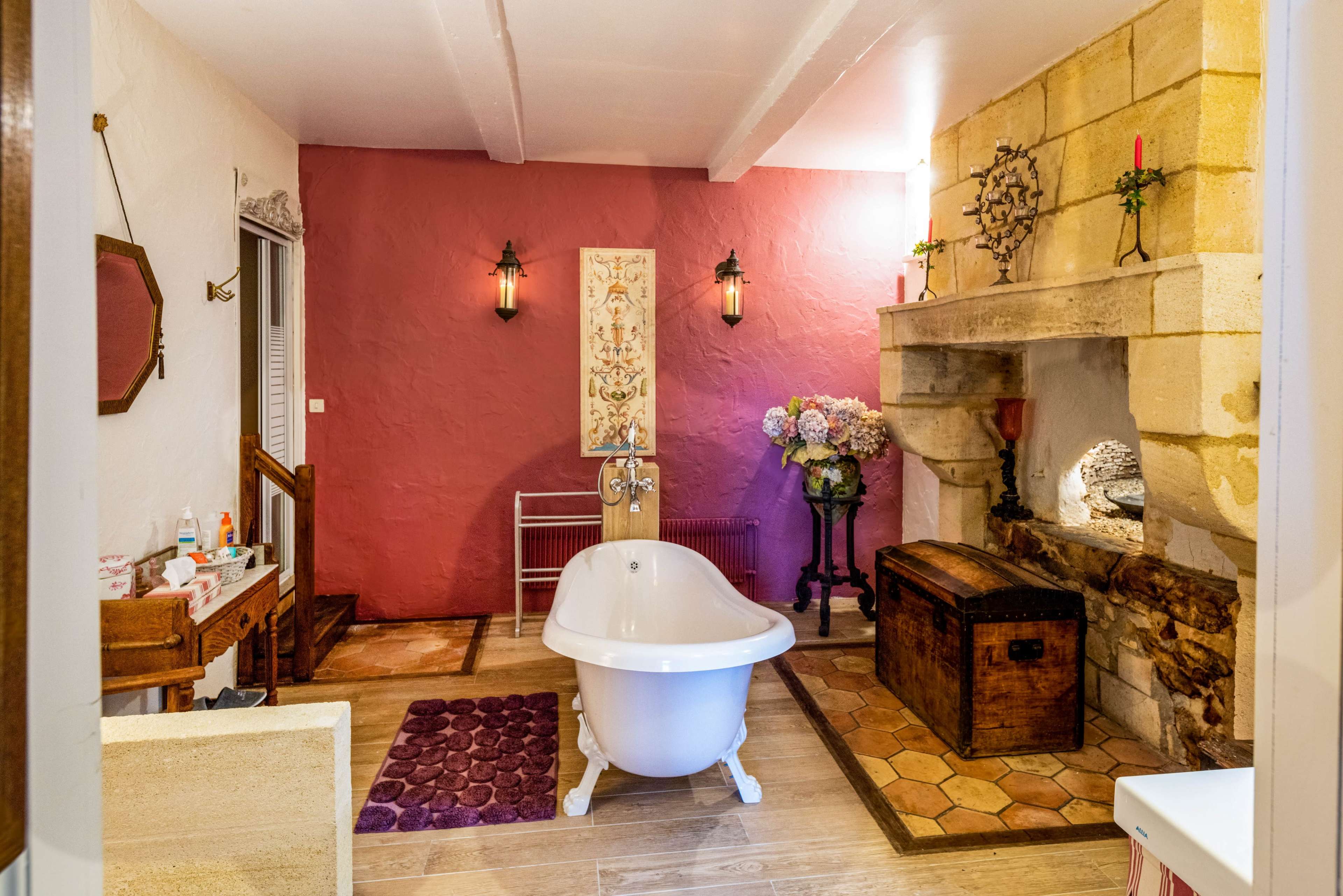 The image shows a bathroom featuring a freestanding white bathtub, a stone fireplace with decorative elements, and a wall painted in a deep pink hue, accented by flowers and candles.