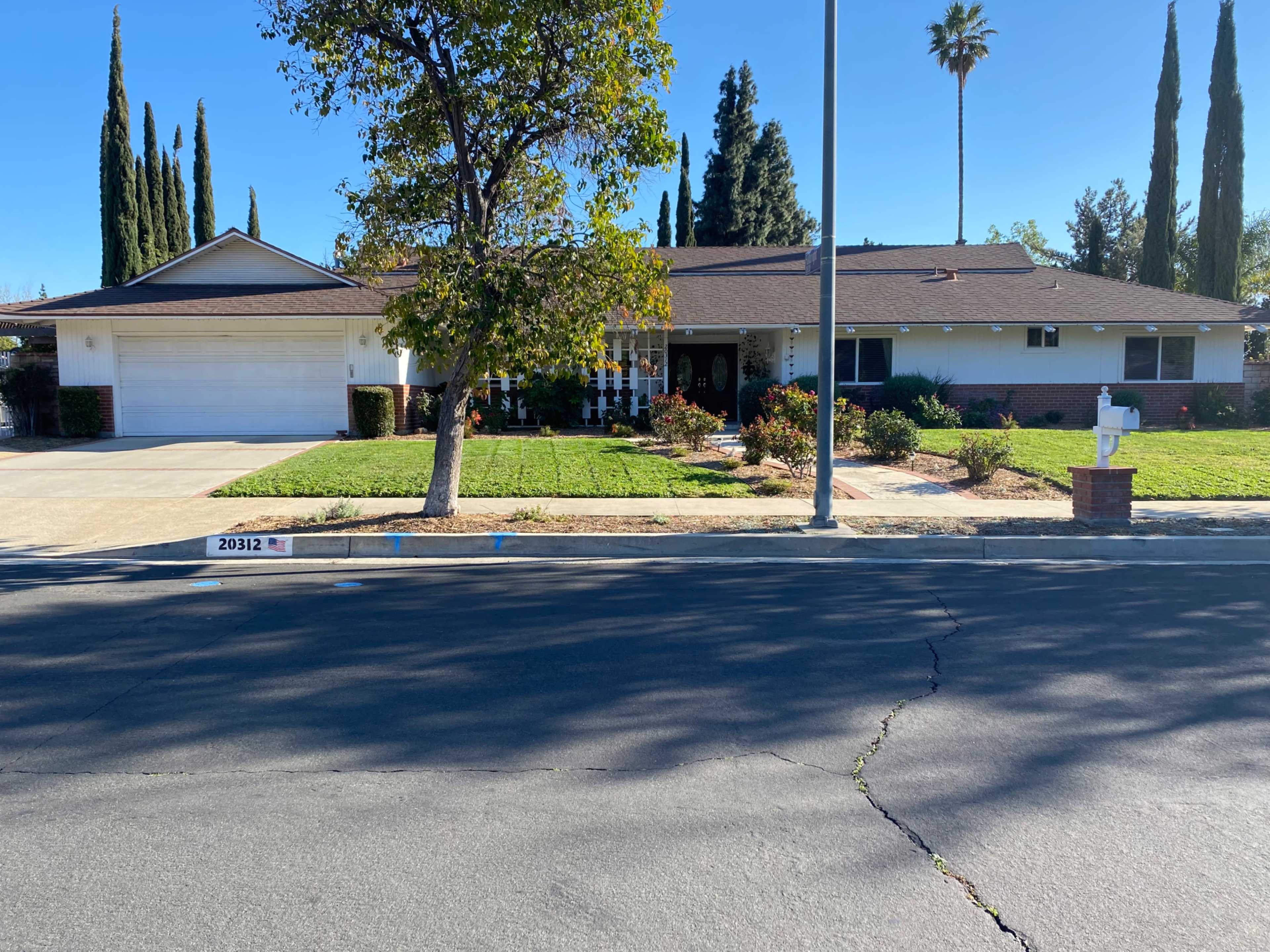 A single-story house with a manicured lawn and tall trees is situated on a quiet residential street.
