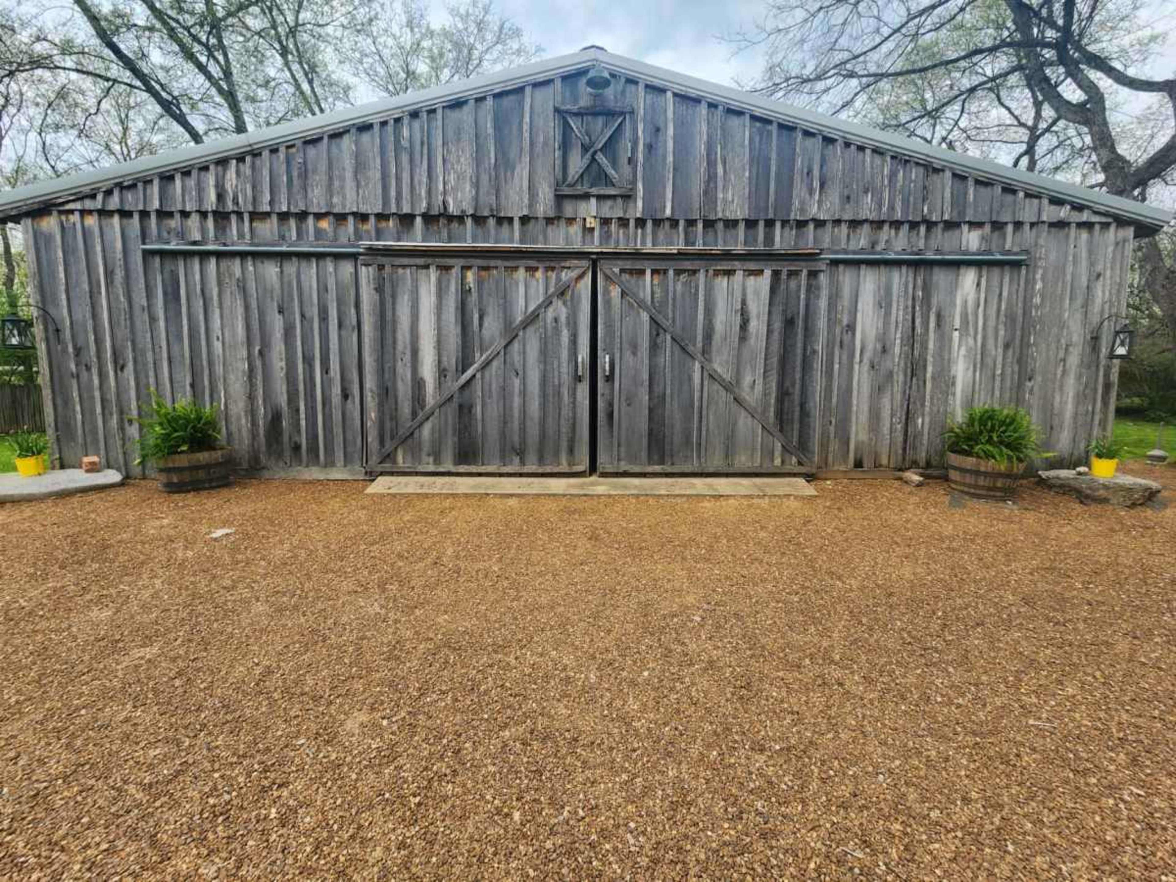 A weathered wooden barn with double doors, situated in a gravel area surrounded by plants in barrels.