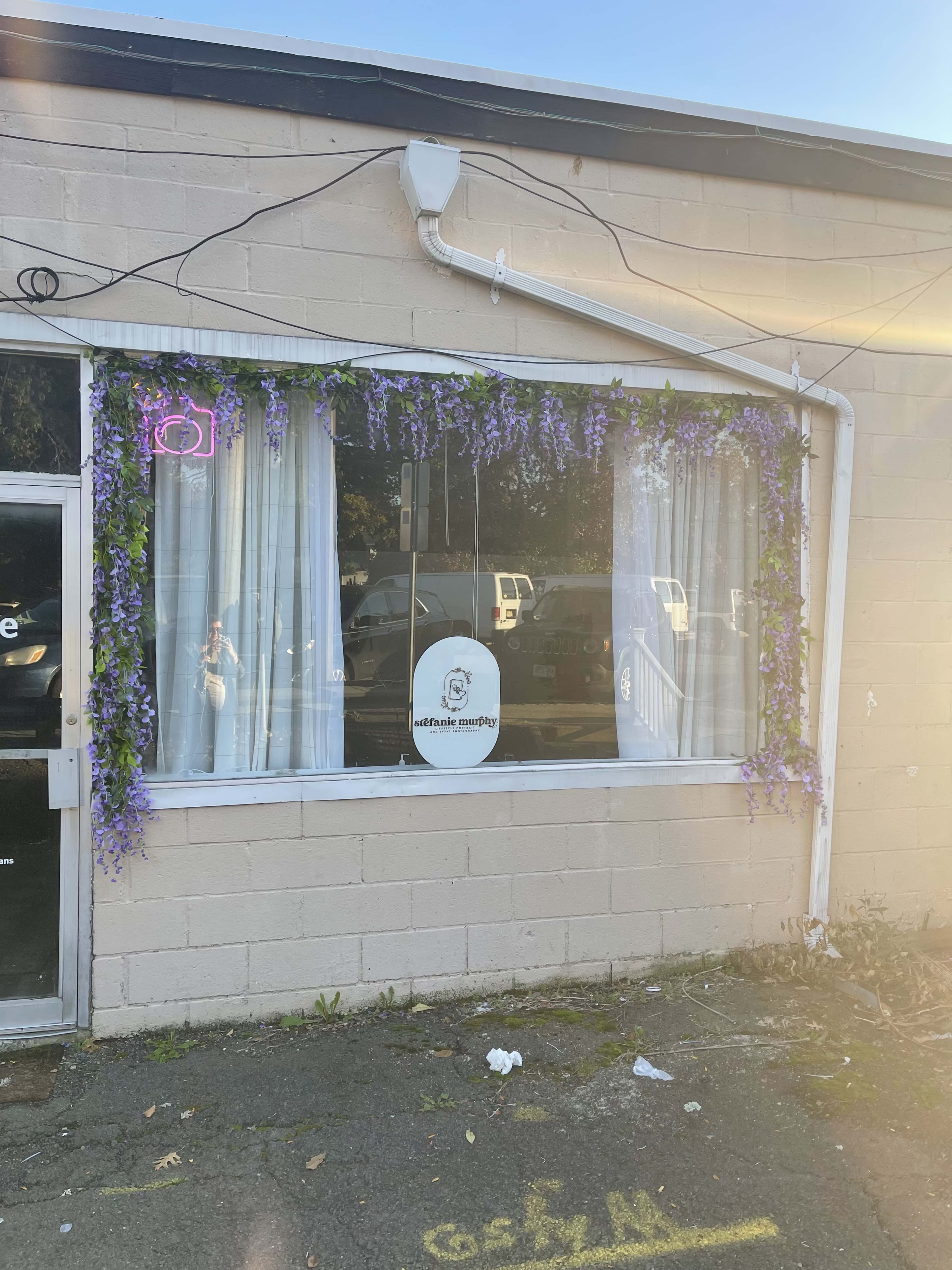 A storefront window adorned with purple floral decorations and sheer white curtains.