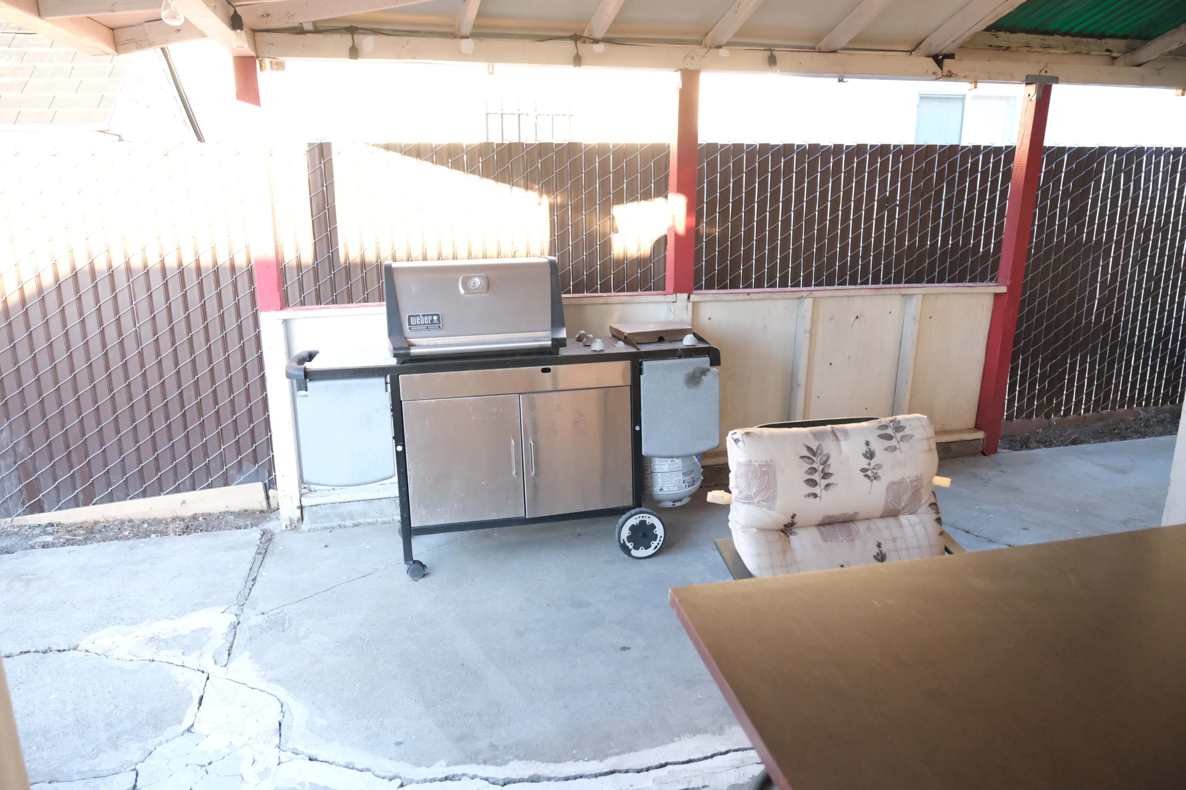 The image shows an outdoor patio with a barbecue grill on a cart next to a cushioned chair and a fenced area.