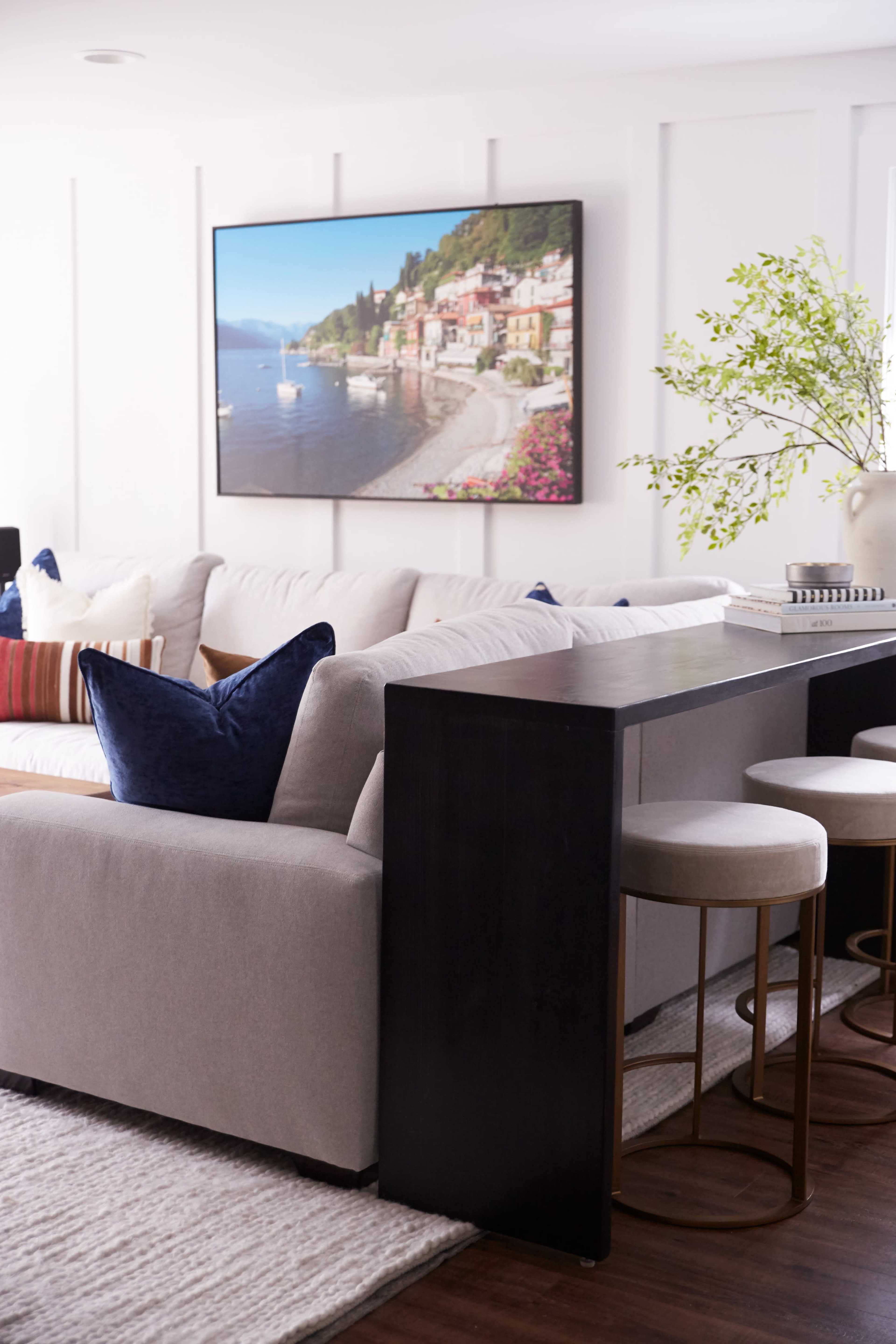 A modern living room with a light-colored sectional sofa, a black bar table with stools, and a large landscape painting hanging on the wall.