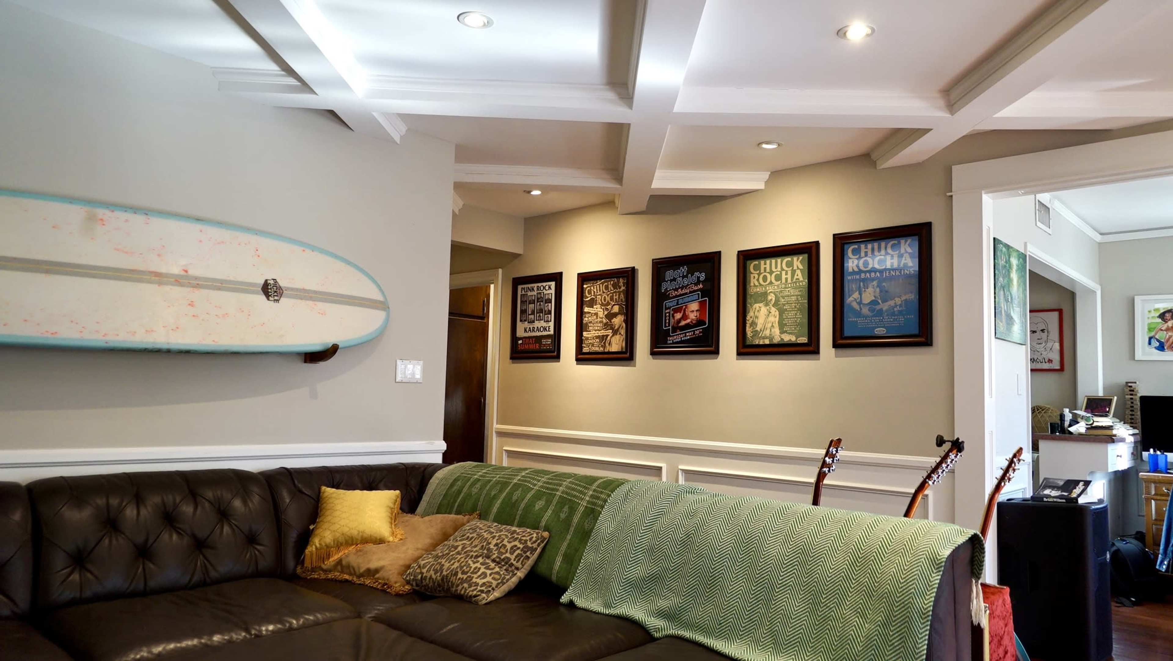 The image shows a living room with a black leather couch, a green throw blanket, a surfboard mounted on the wall, and several framed posters hanging on the wall.