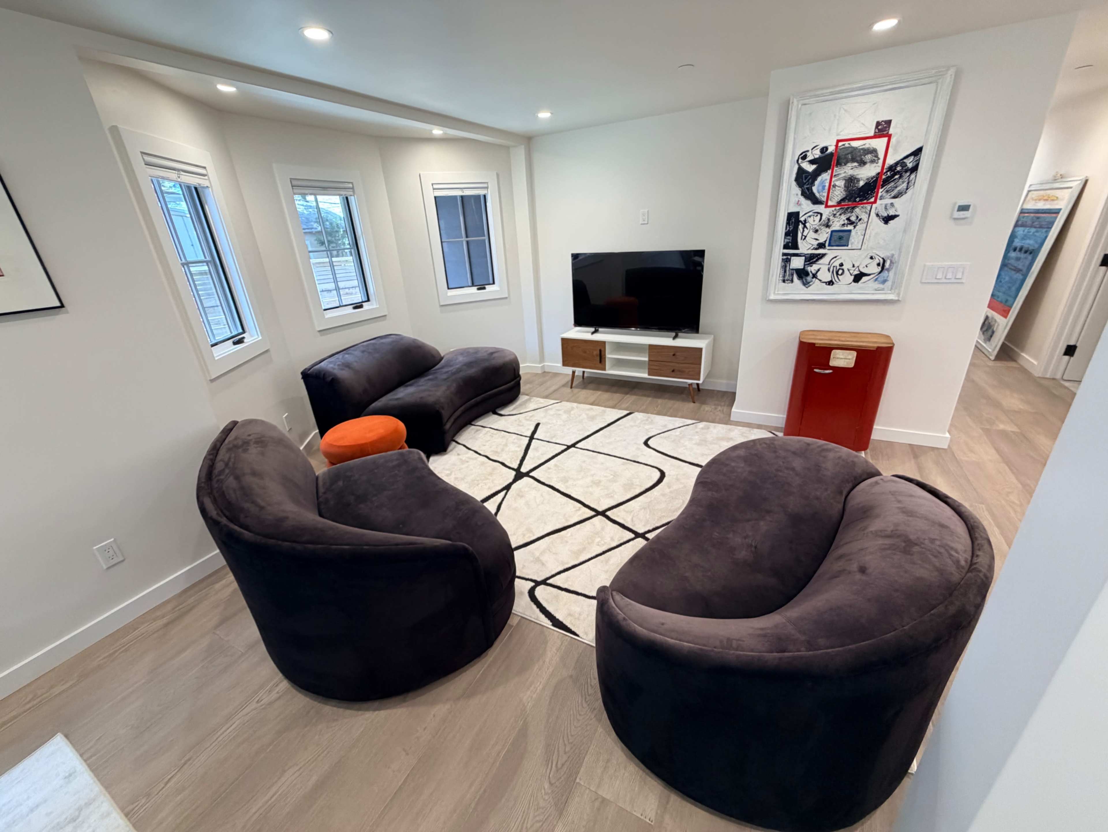 The image shows a modern living room with two dark, plush chairs, a circular orange ottoman, a white rug with a black pattern, and a wooden TV stand against a white wall.