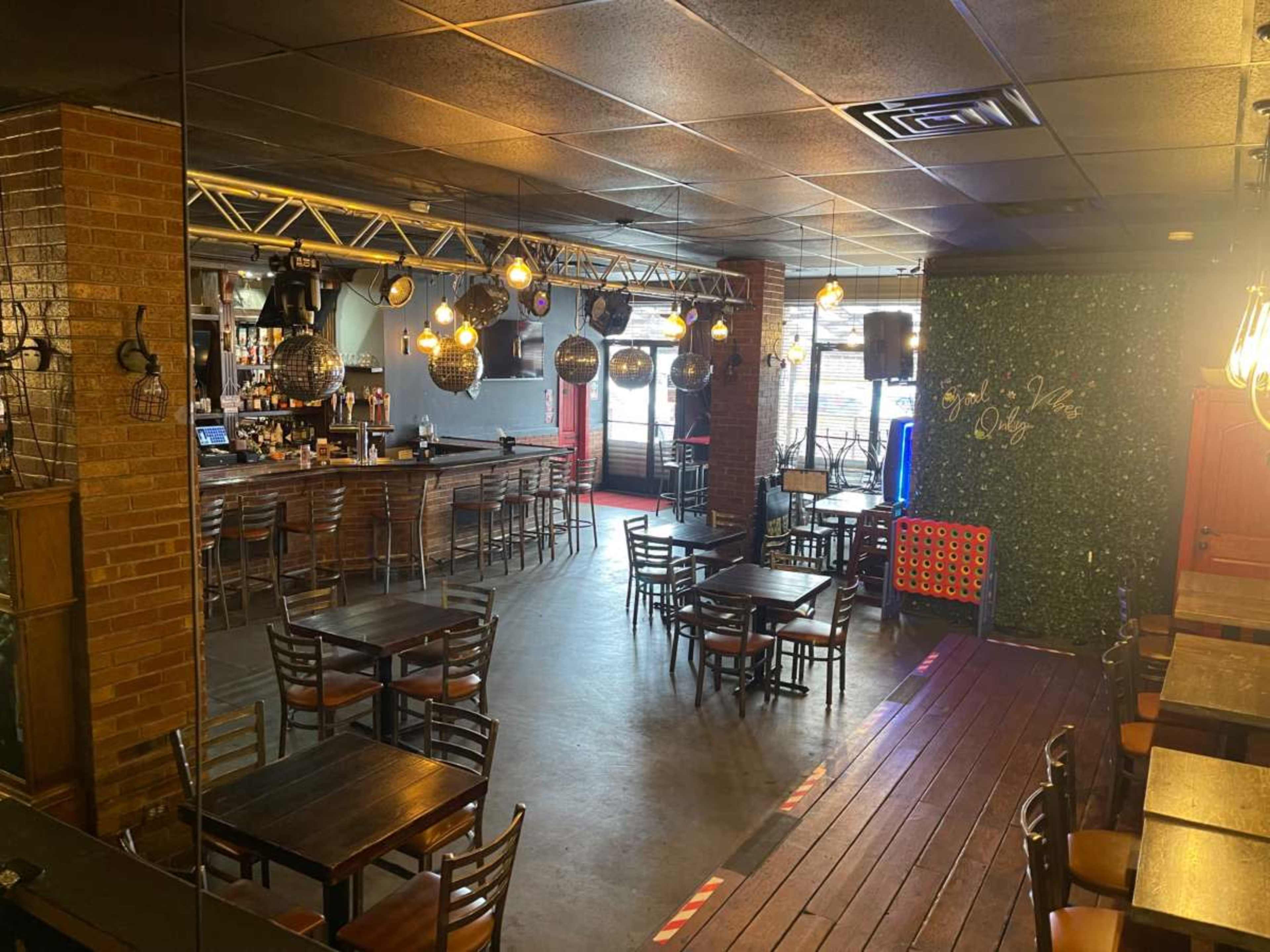 The image shows a dimly lit bar and dining area with wooden tables, chairs, and a bar in the background, featuring hanging lights and decorations on the walls.