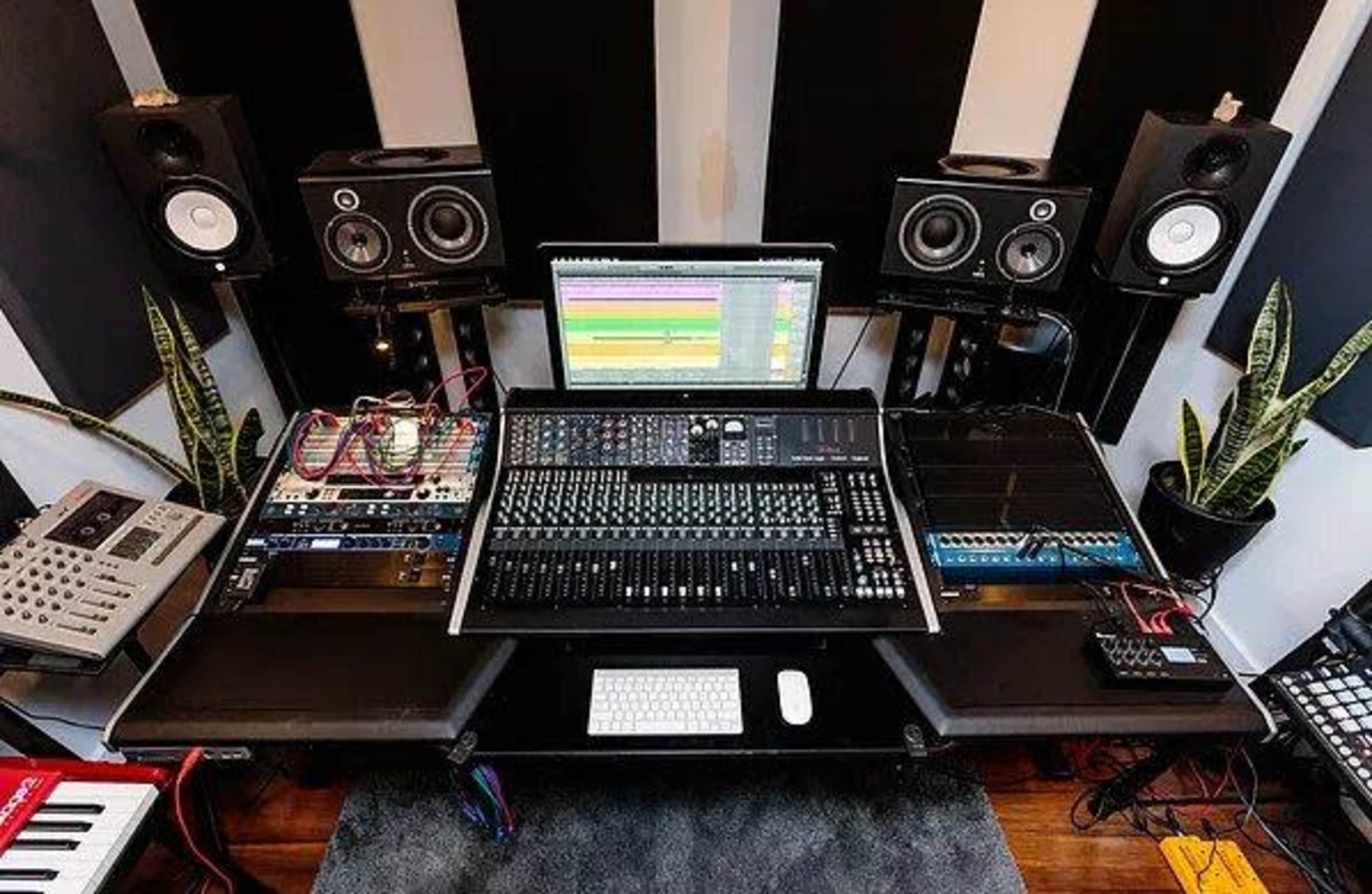 A music production workstation with a large mixing console, computer, speakers, and various audio equipment arranged around it.